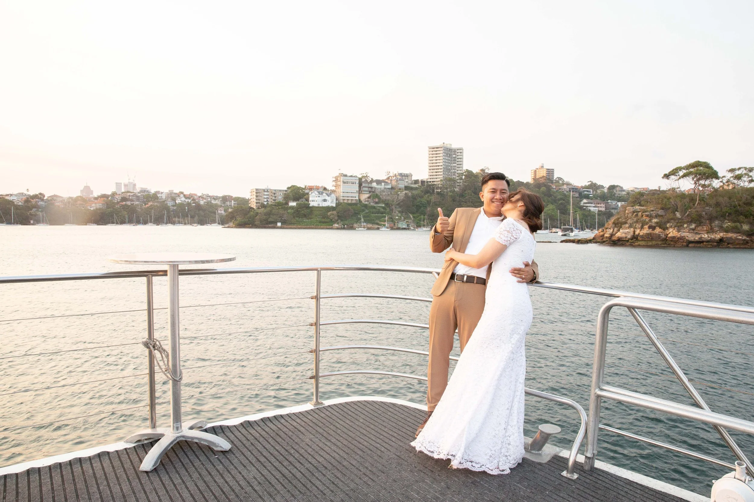 A couple in wedding attire hugging on a boat deck near a body of water with a cityscape and trees in the background.