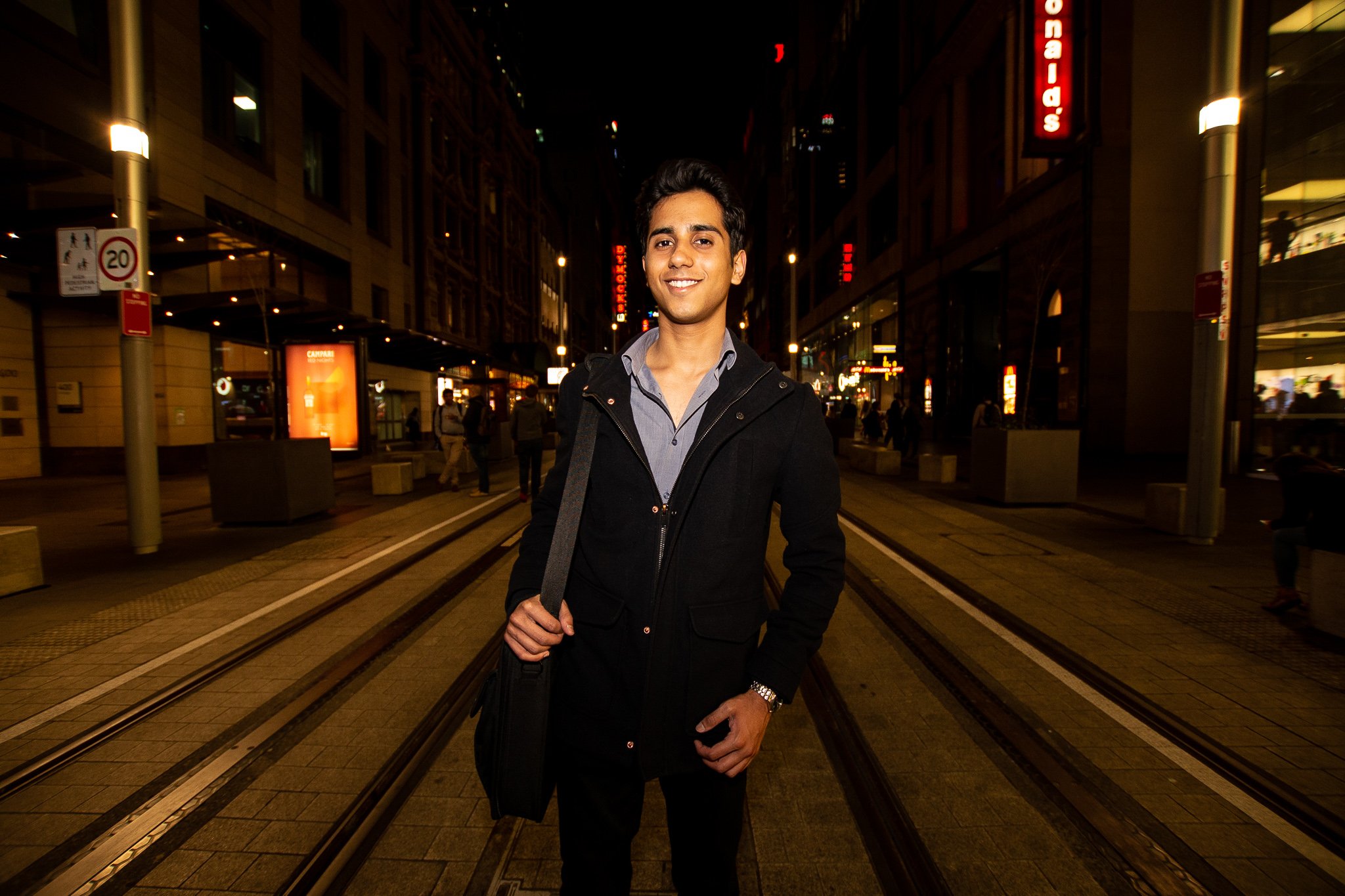 A young man standing on a city street at night, smiling at the camera, with illuminated buildings and signs behind him.