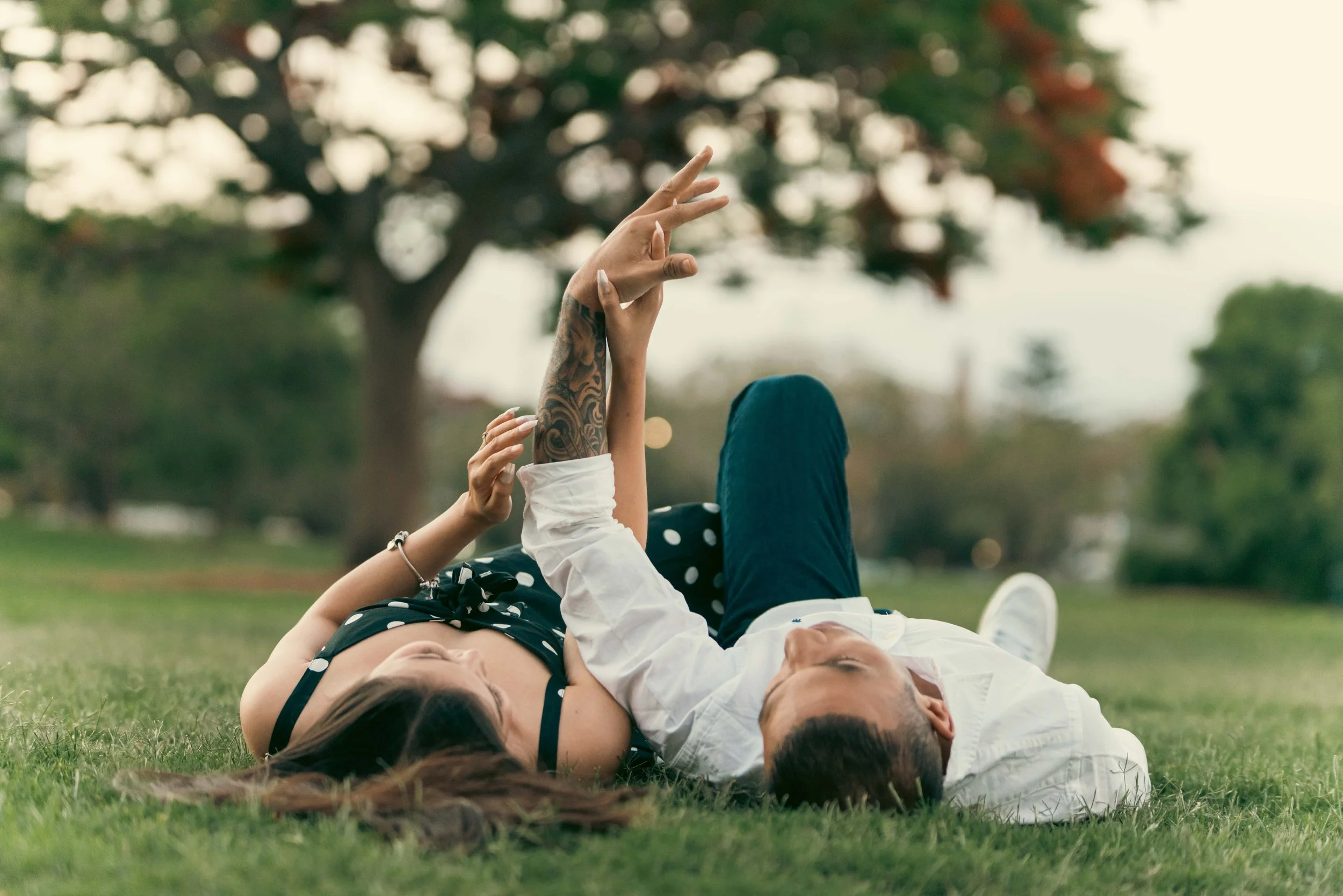A couple lying on the grass in a park, holding hands, with one person showing a tattooed arm. They appear to be relaxed and enjoying each other's company.