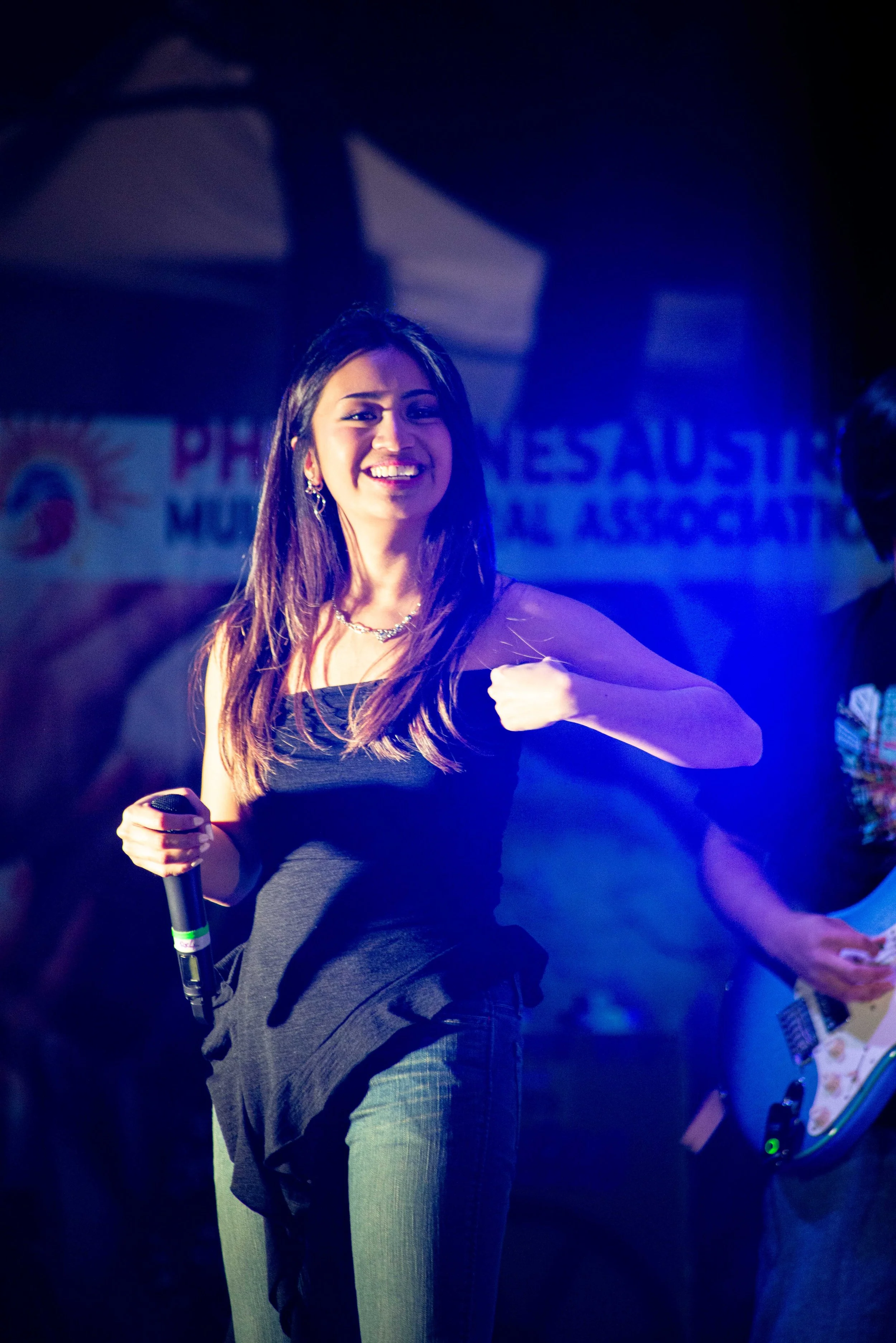 A woman performing on stage, holding a microphone, smiling, with a dark background and a blurred banner in the background.