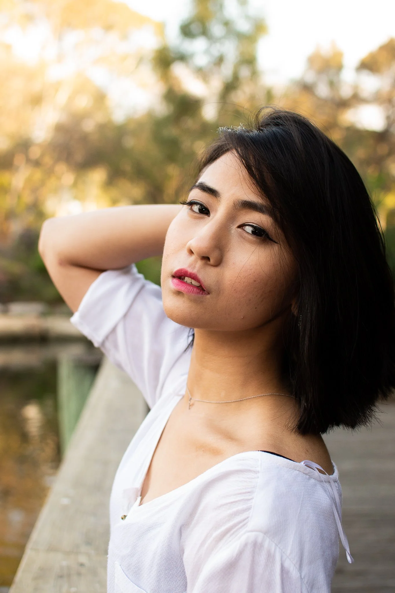 A young woman with shoulder-length black hair posing outdoors with trees in the background during golden hour, wearing a white top and a delicate necklace.