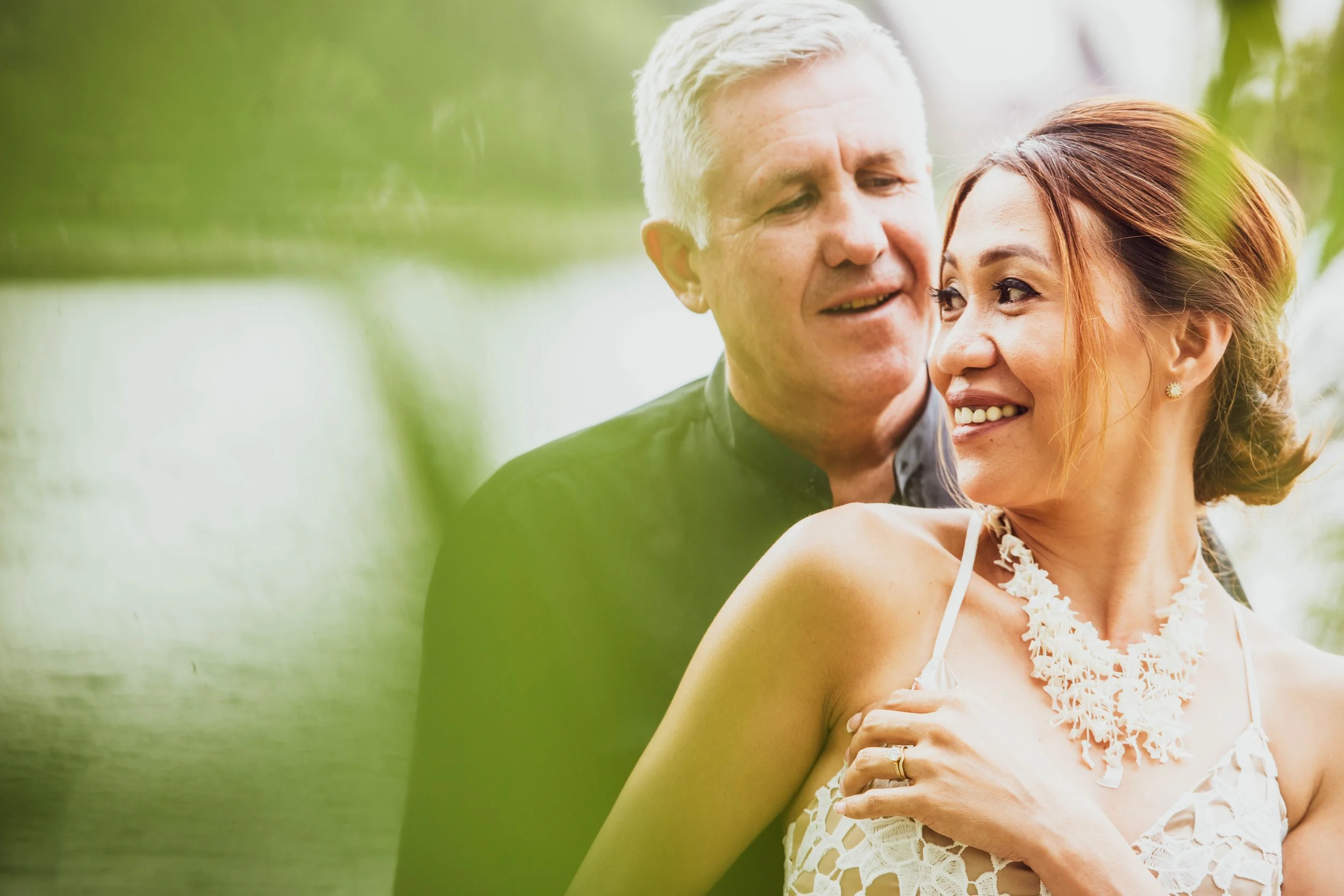 A smiling woman in a white lace dress and decorative necklace, with short brown hair, in front of an older man with light-colored hair, close to her in an outdoor setting with blurry green foliage and a body of water in the background.