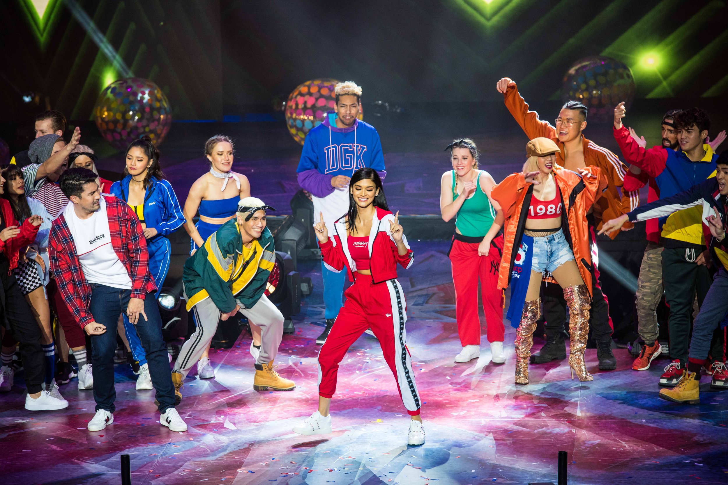 A lively group of young people dancing and celebrating on a stage with colorful lighting and disco balls.