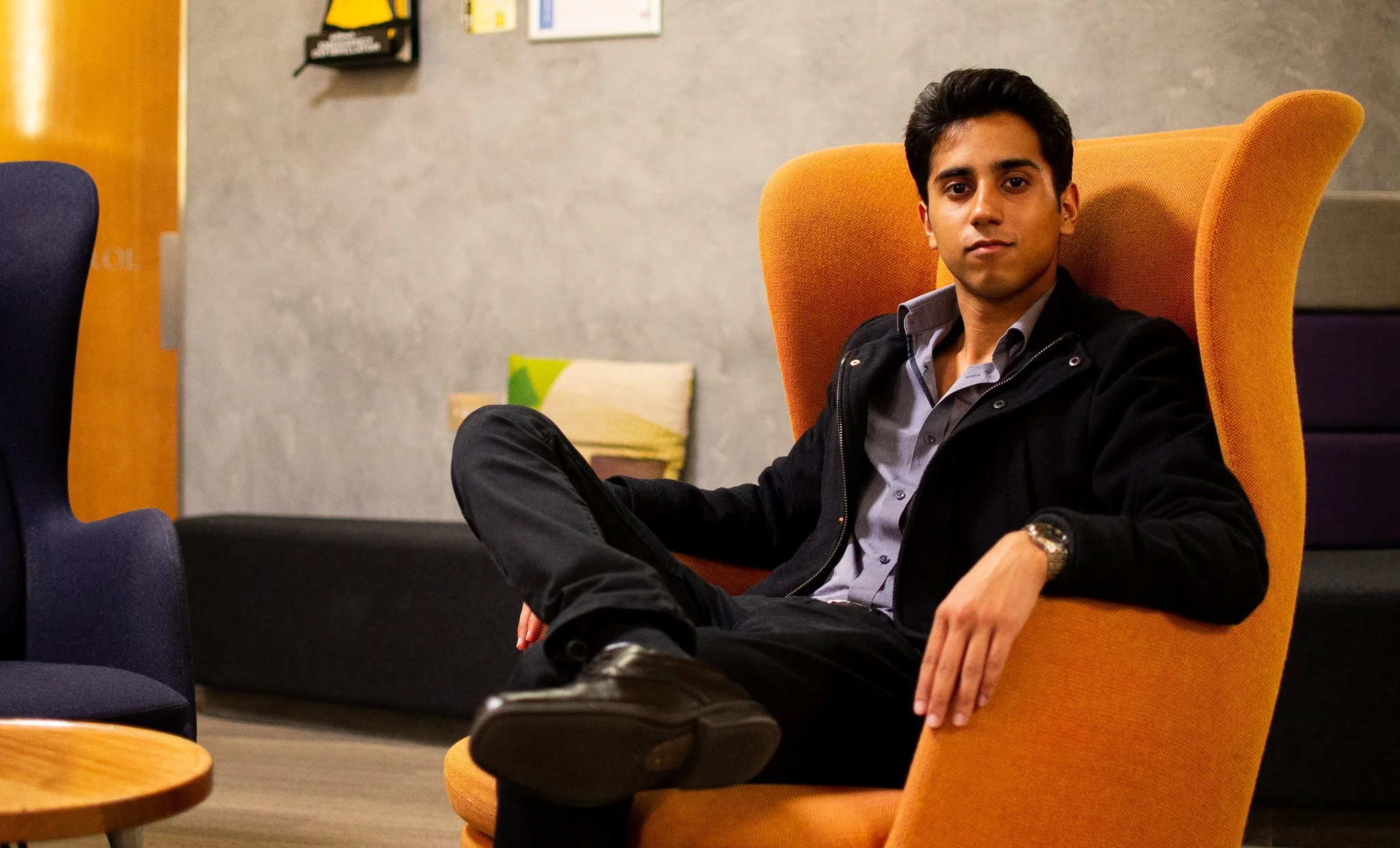 A young man sitting in an orange wingback chair, wearing a black jacket, light gray shirt, and black pants, with his legs crossed and leaning back with his left arm resting on the armrest, in a modern indoor setting.
