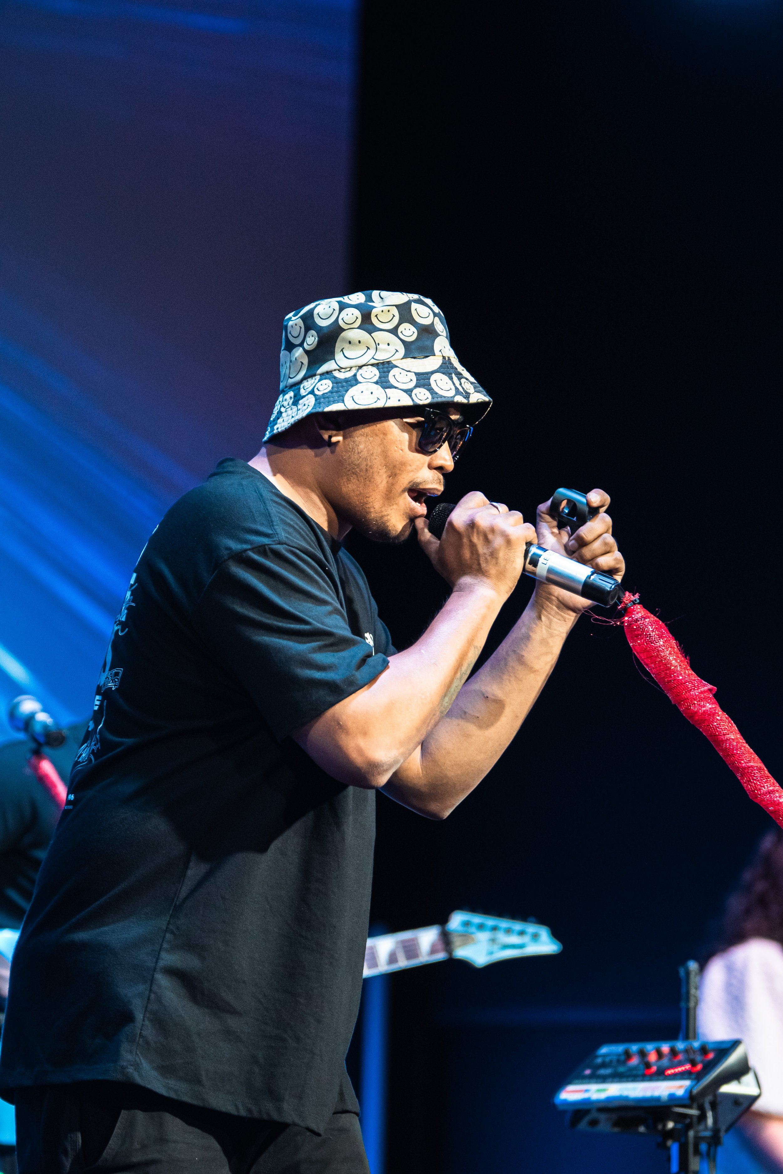 A male performer wearing black sunglasses and a bucket hat with smiley faces, singing into a microphone on stage with musical equipment around him.