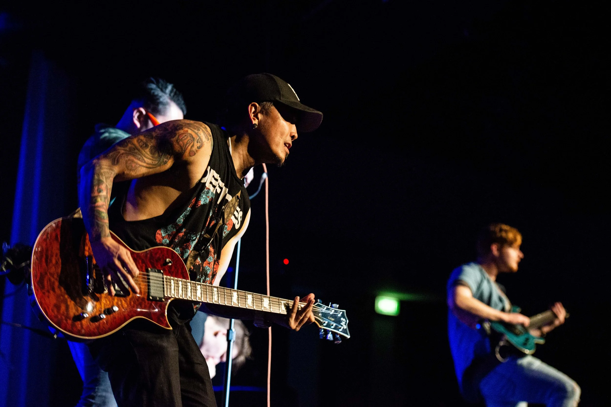 Musicians performing on stage at a concert, with one playing an electric guitar and another playing a small guitar, both wearing casual clothing and cap, against a dark background.