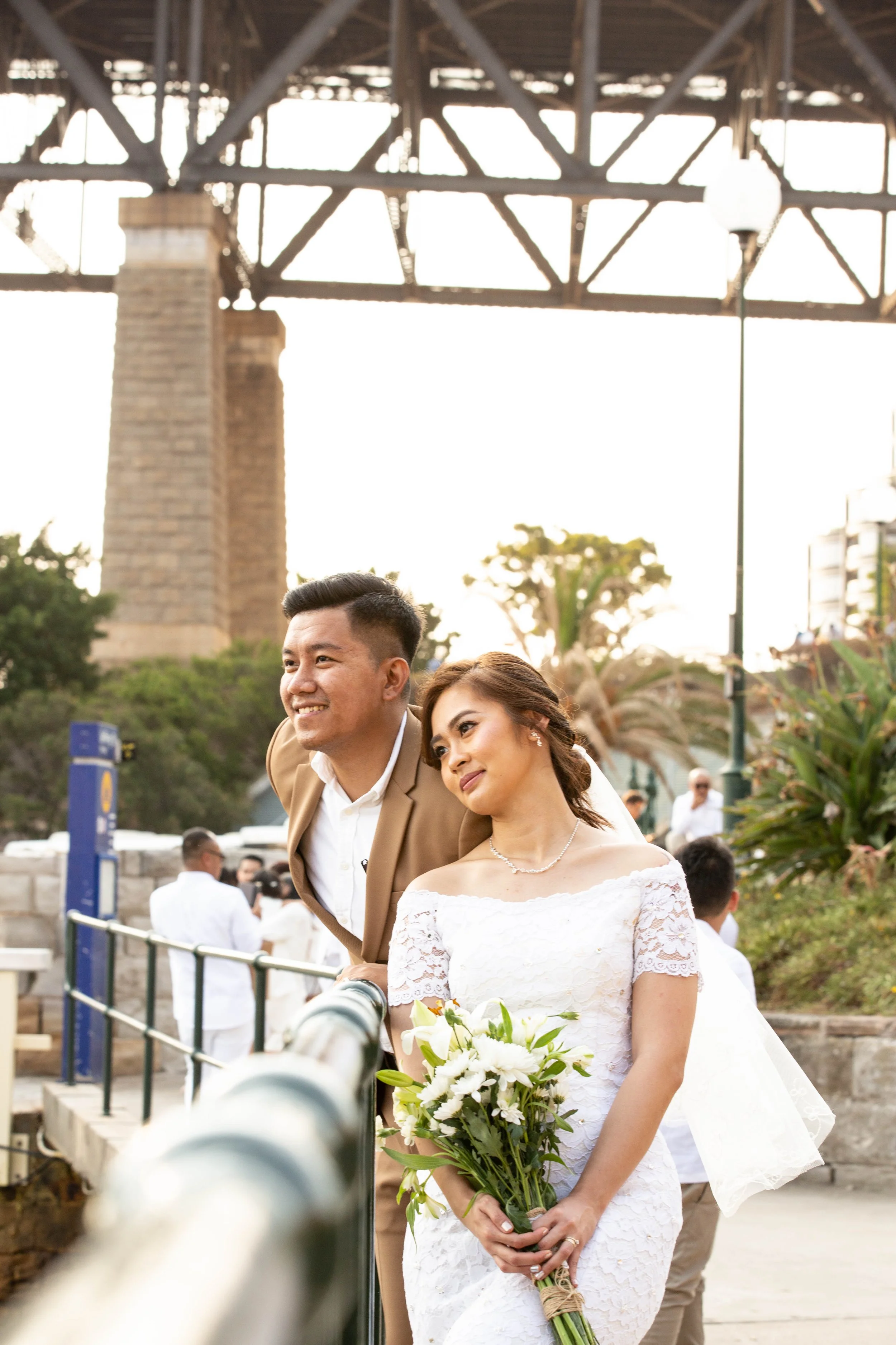 A bride holding a bouquet of white flowers stands next to a smiling man at a wedding or outdoor event, with the San Diego-Coronado Bridge in the background.