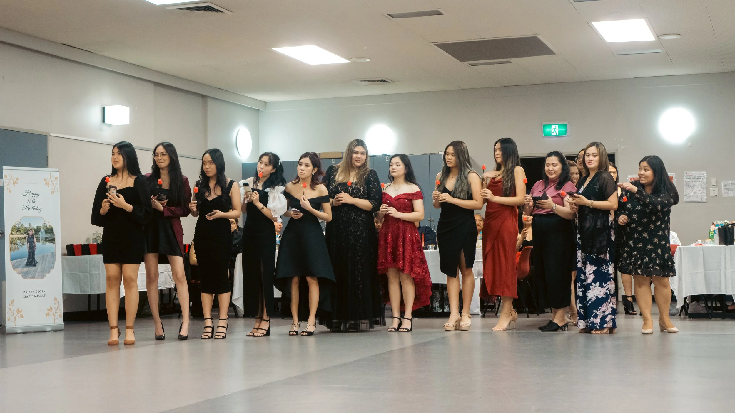 Group of women standing in a line at a celebration, holding small red and black objects, dressed in formal attire, in a room decorated for a birthday party.