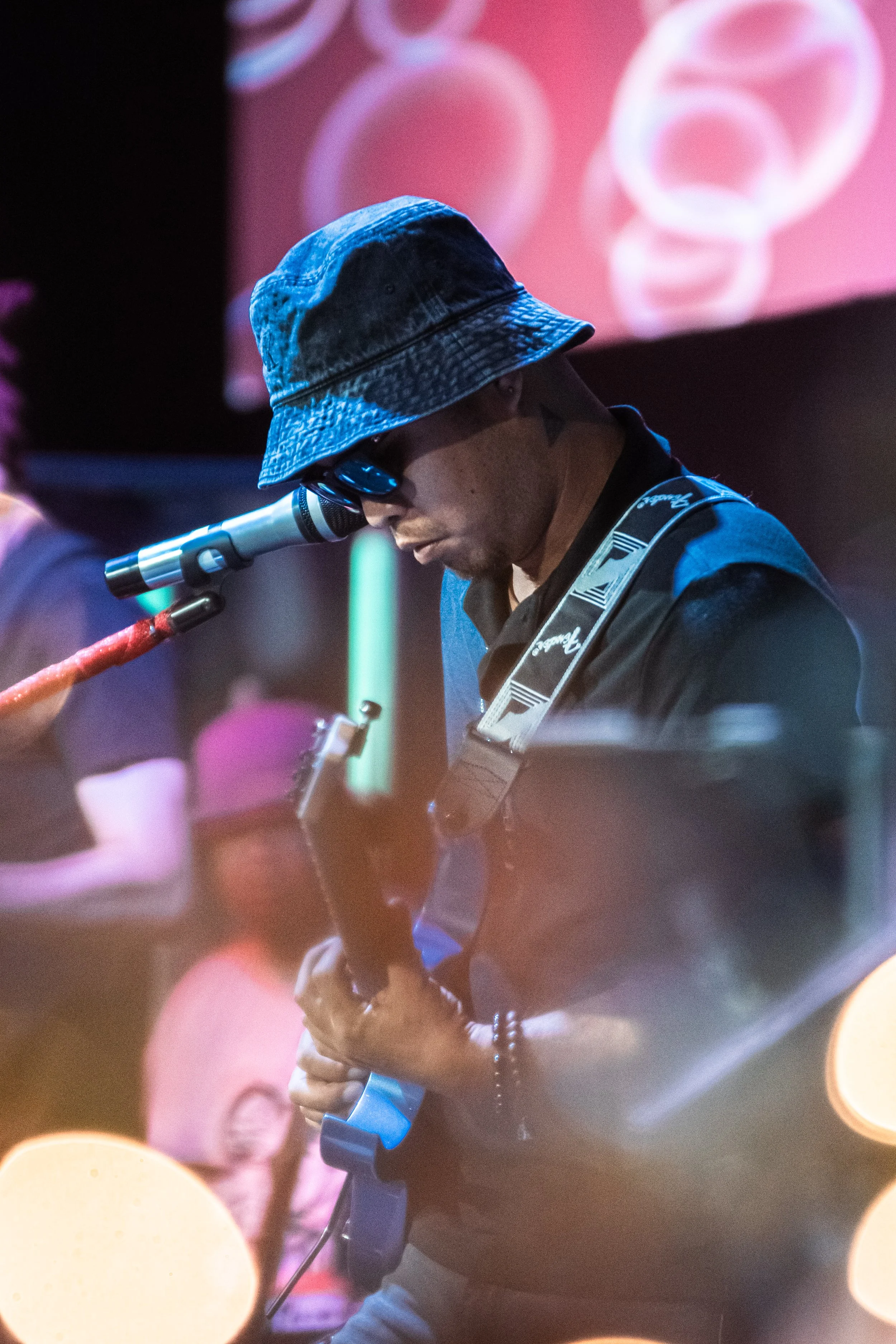 A male musician wearing a blue bucket hat and sunglasses playing an electric guitar on stage with colorful lights and blurred background.