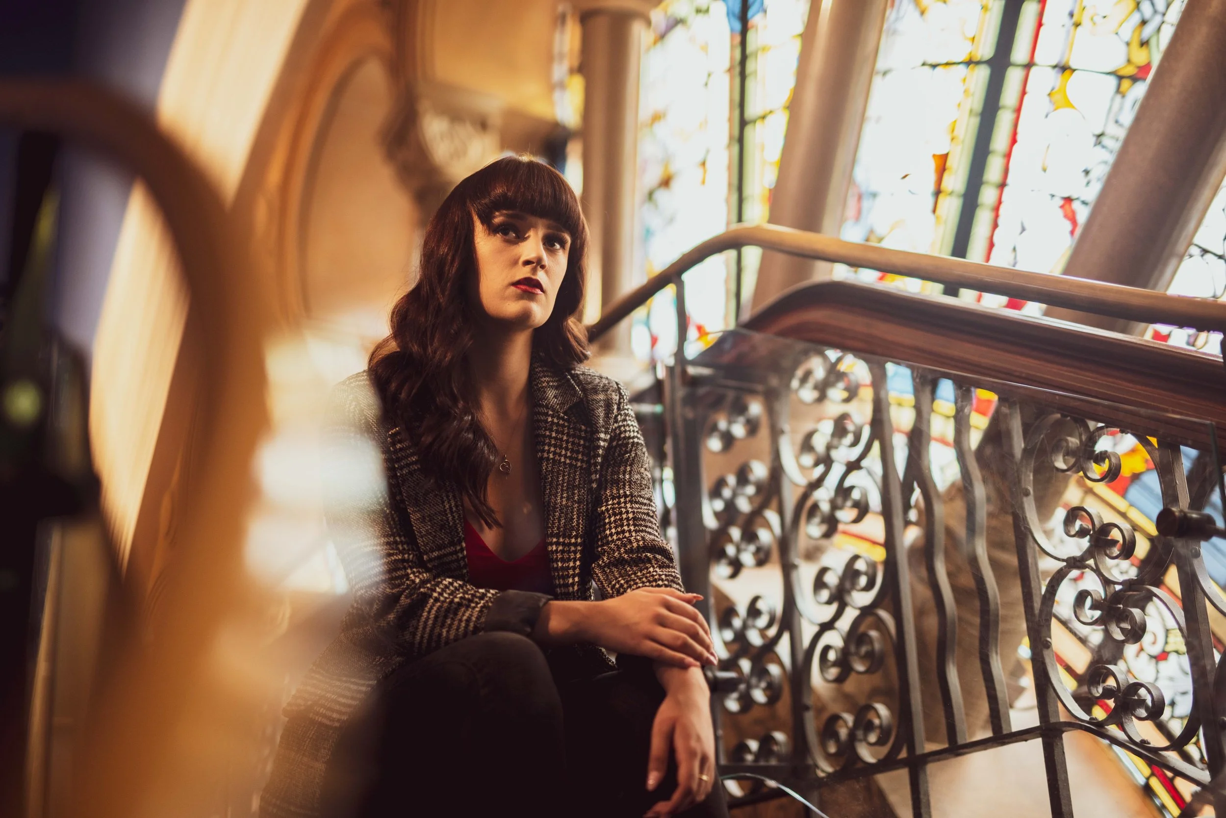 A woman with dark wavy hair and bangs sitting on a staircase next to stained glass windows, wearing a plaid blazer and dark pants, looking contemplative.