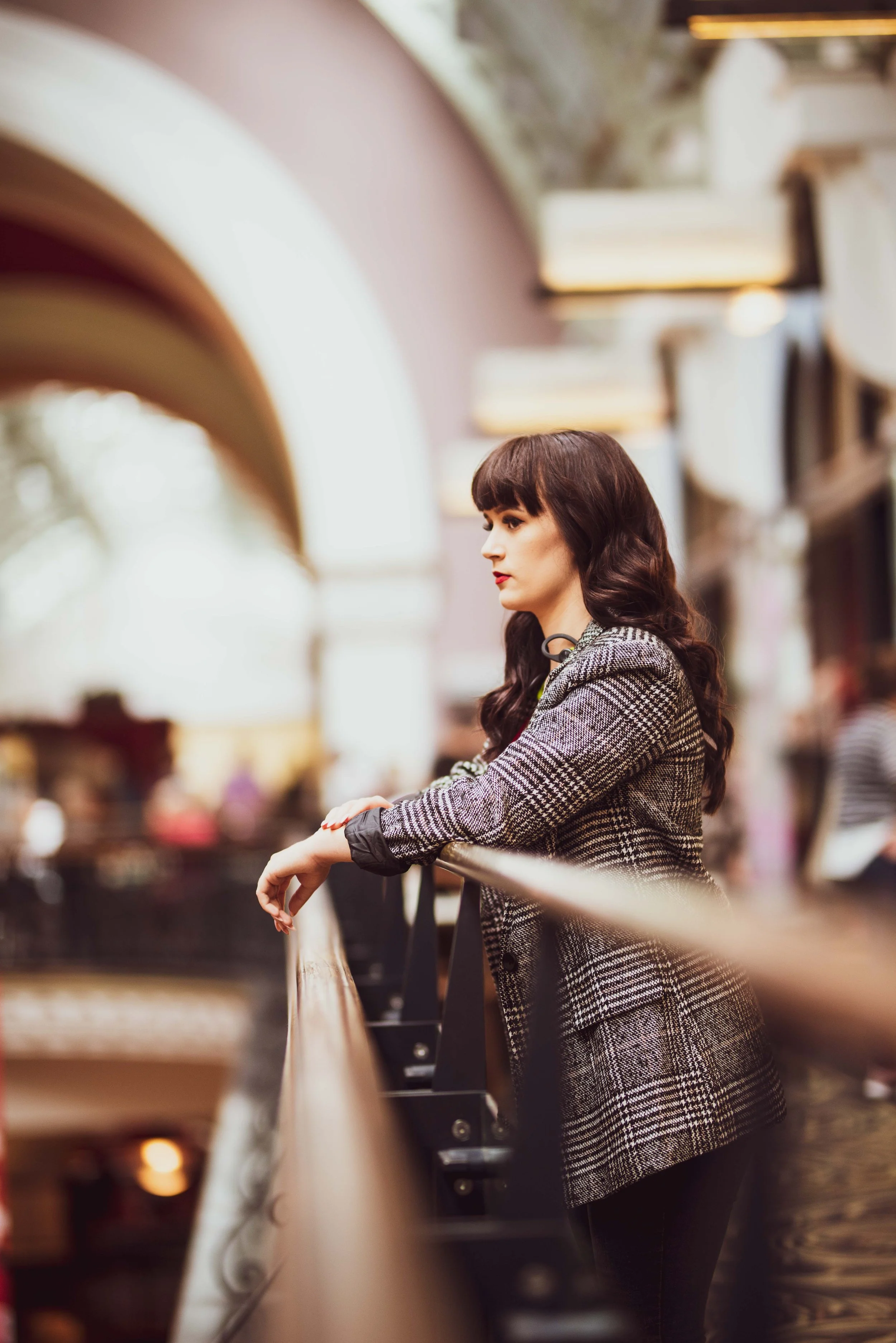 A woman with dark wavy hair and wearing a plaid blazer stands by a railing inside a mall or shopping center.