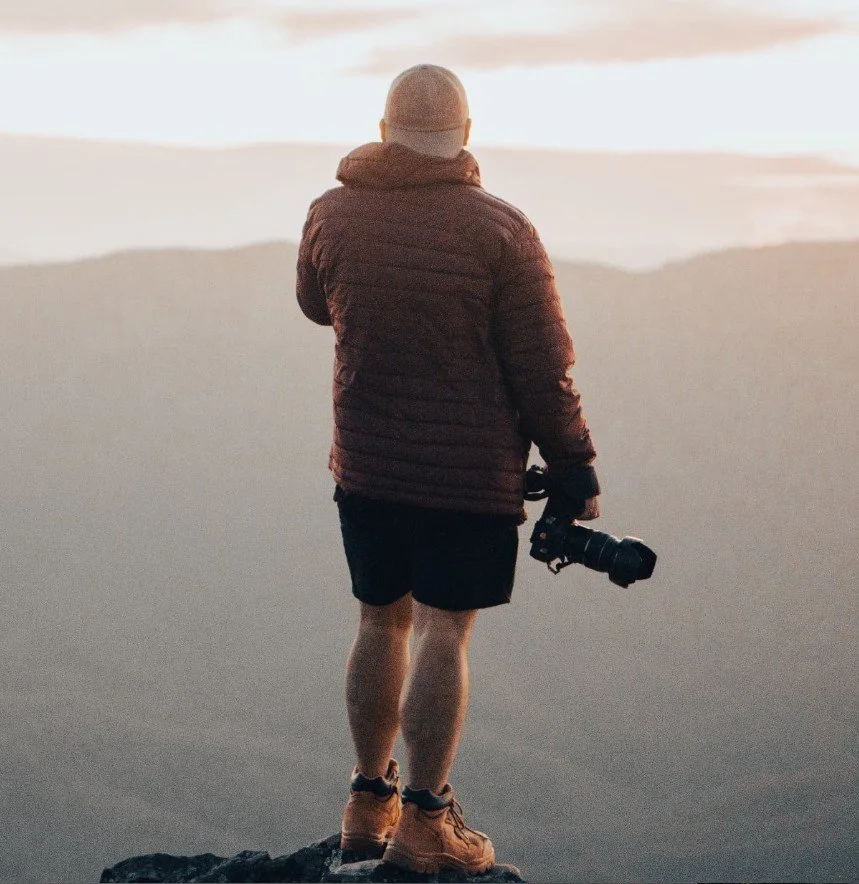Person standing on a mountain or hilltop during sunrise or sunset, holding a camera and looking at a scenic view of mountains and clouds.