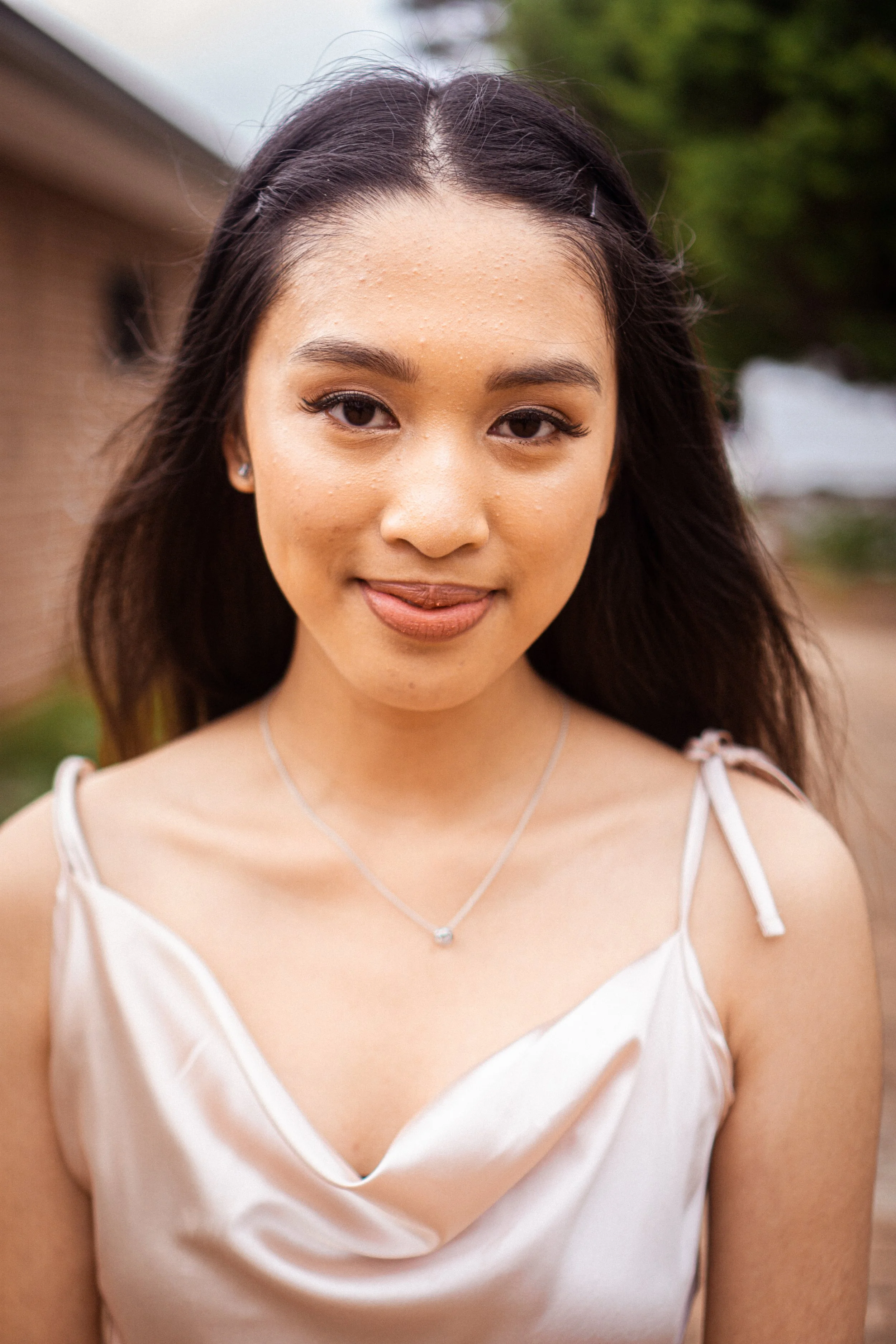 A young woman with long dark hair, wearing a cream-colored satin dress with spaghetti straps, standing outdoors with a blurred background of a brick building and trees.