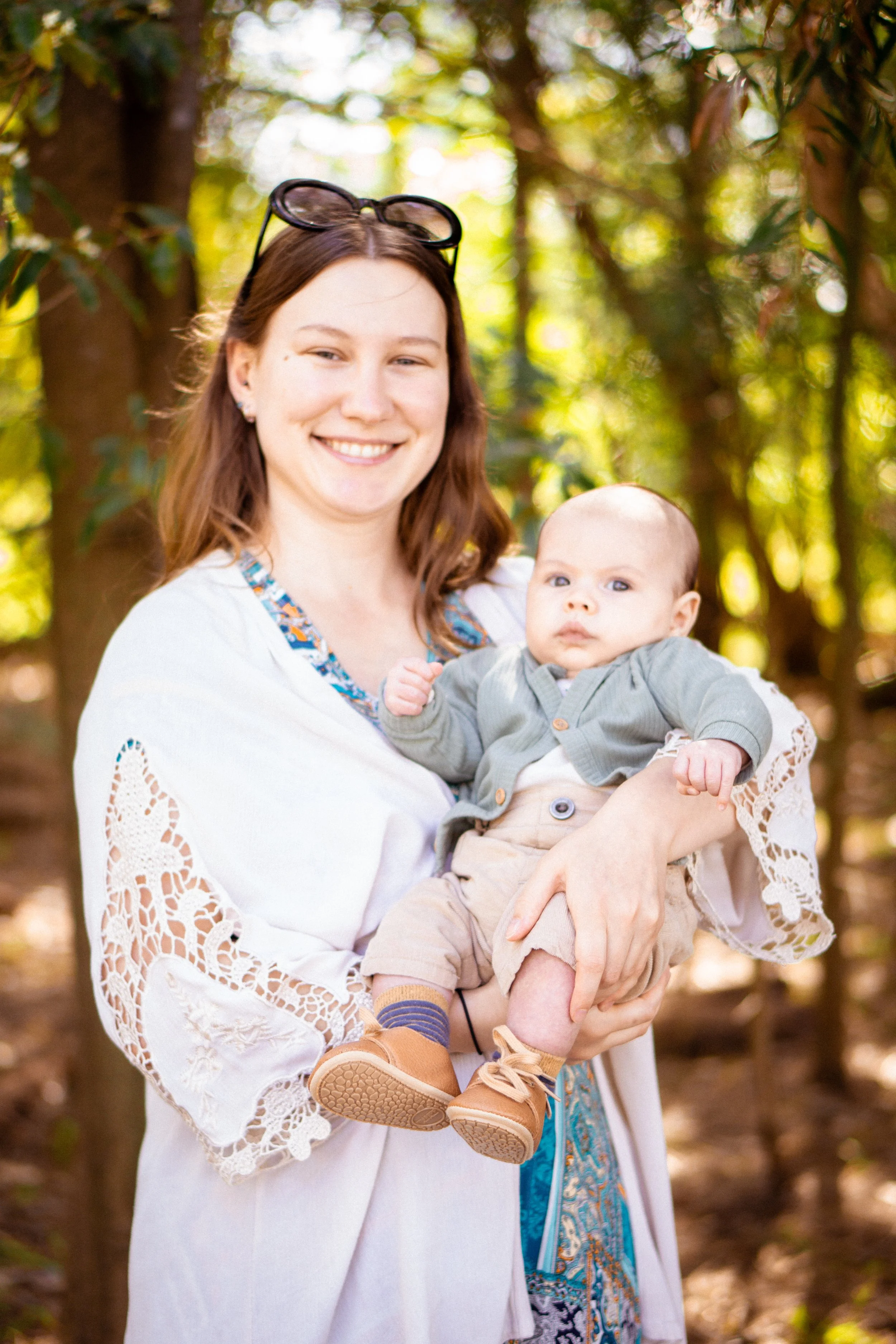 A young woman with sunglasses on her head smiling while holding a baby boy outdoors in a wooded area during daytime.