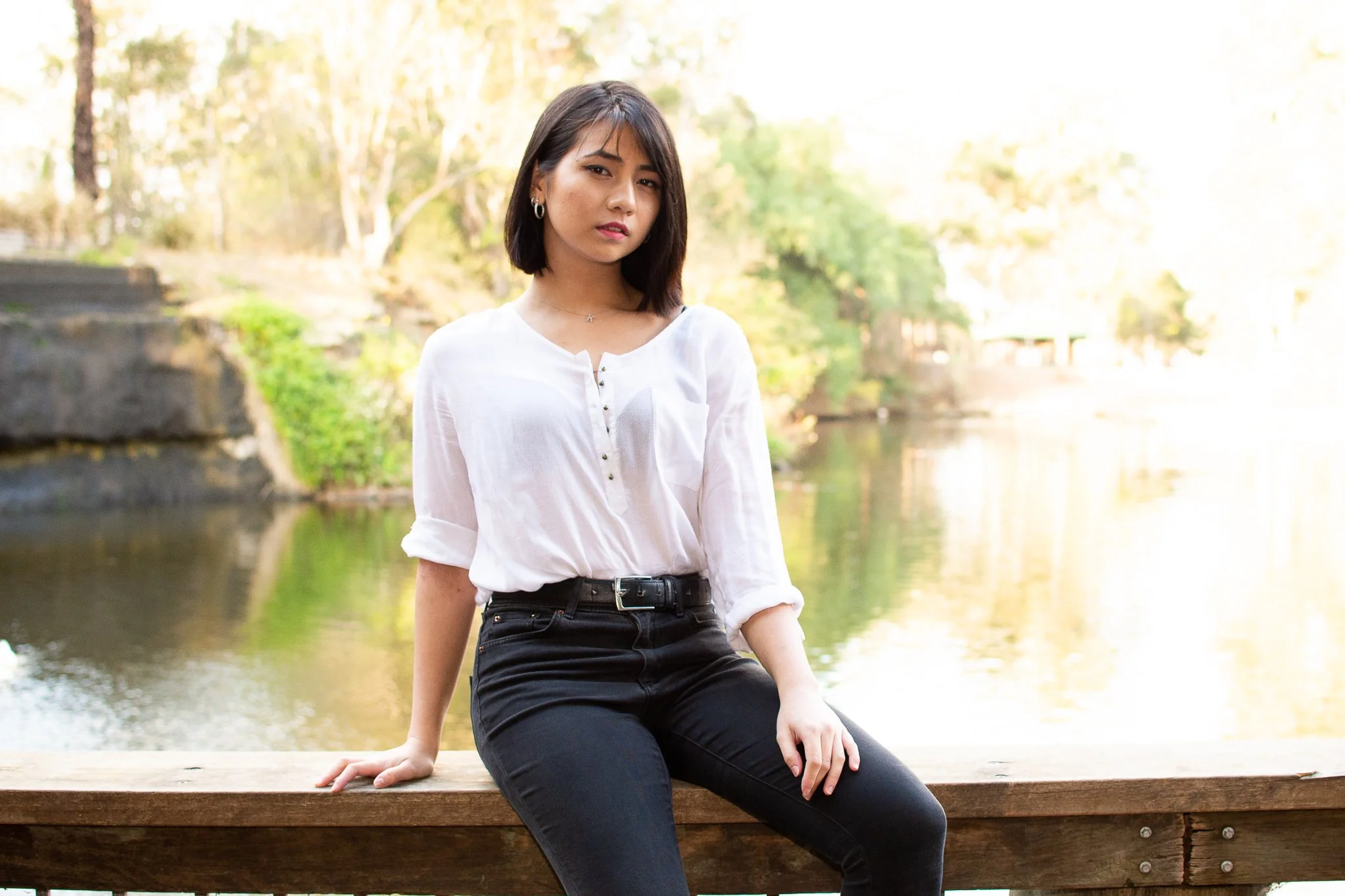 A young woman with shoulder-length dark hair sits on a wooden railing near a body of water, with trees and foliage in the background.