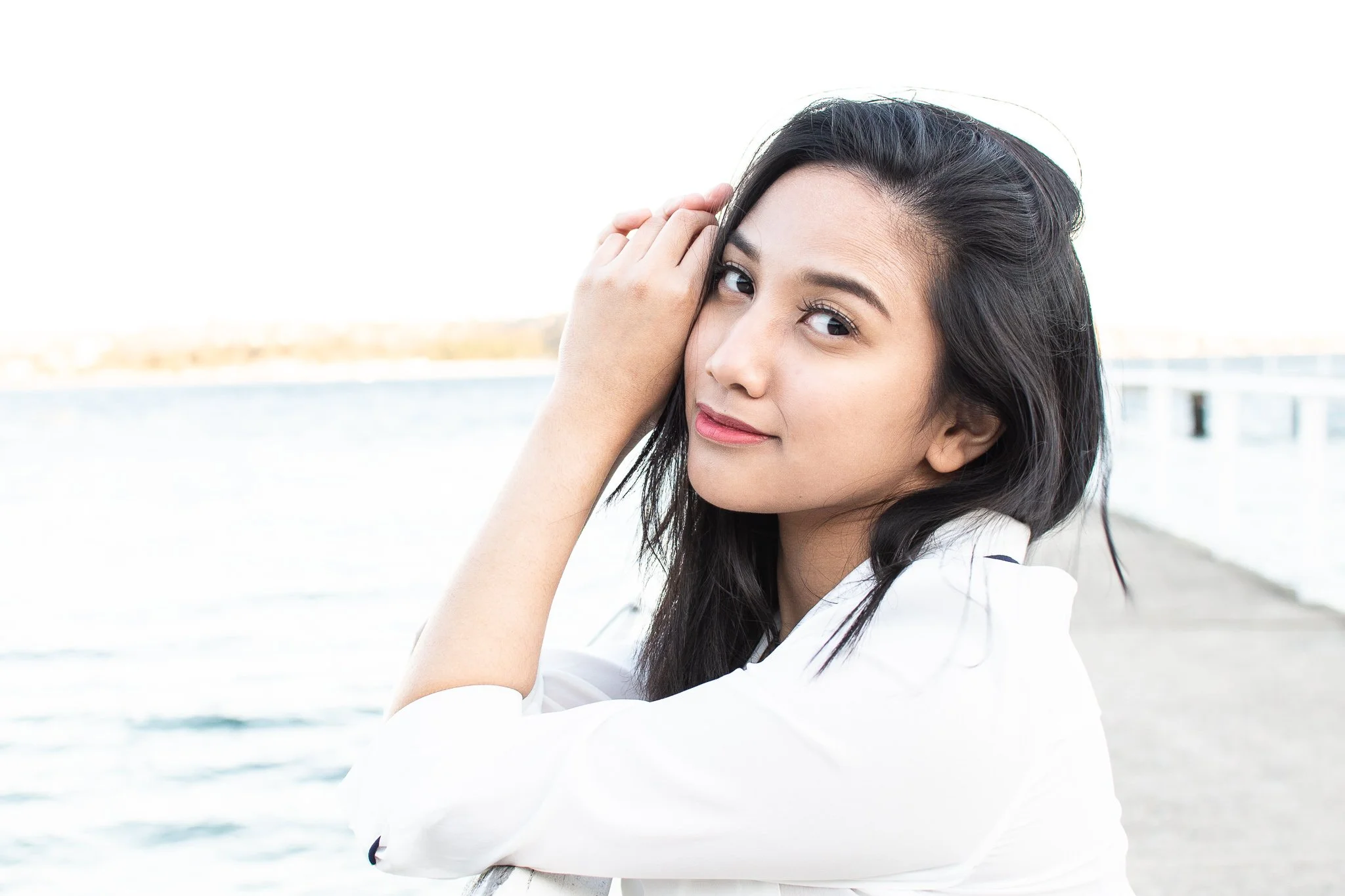 A young woman with dark hair is smiling and posing outdoors near the water, with her hand resting on her head.