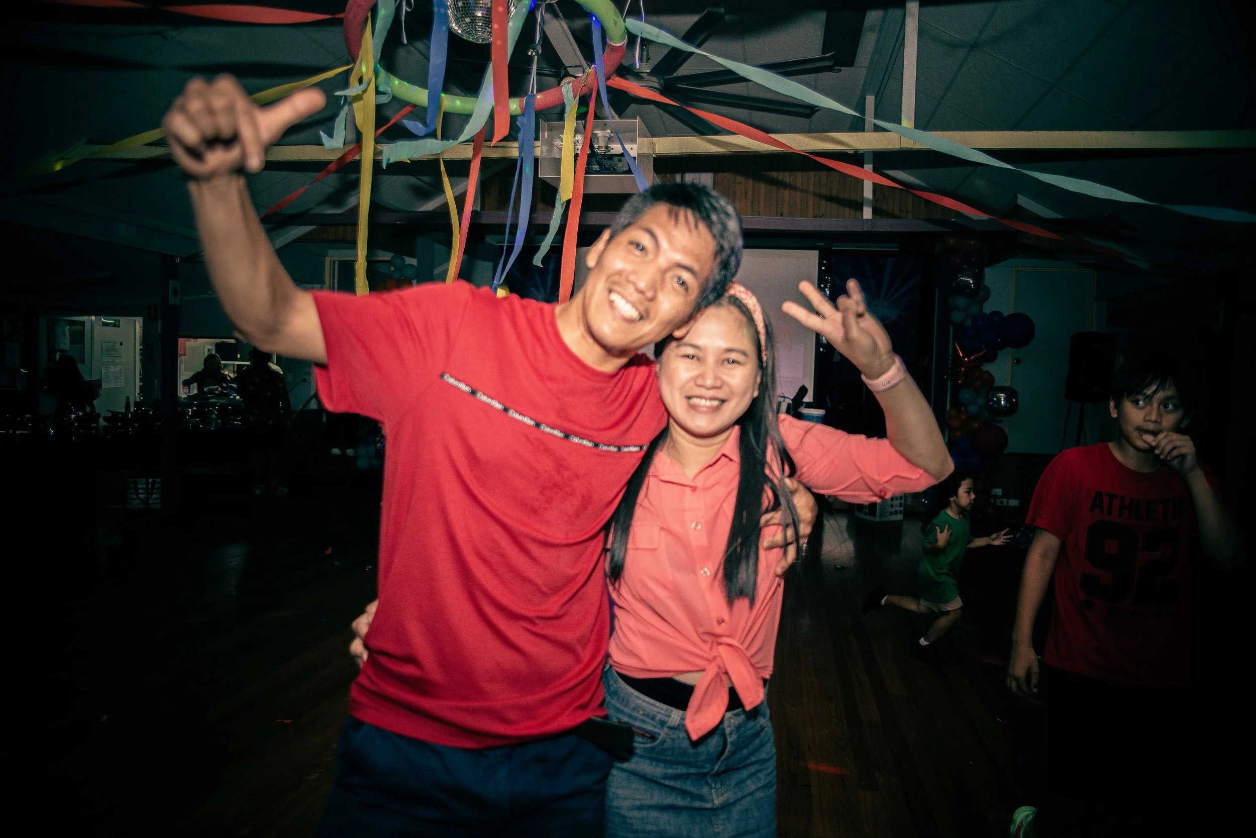 A man and woman with light skin smiling, posing together at a party. The man is wearing a red t-shirt and the woman is in a pink button-up shirt tied at the waist. They are surrounded by colorful ribbons hanging from the ceiling, and children are pla