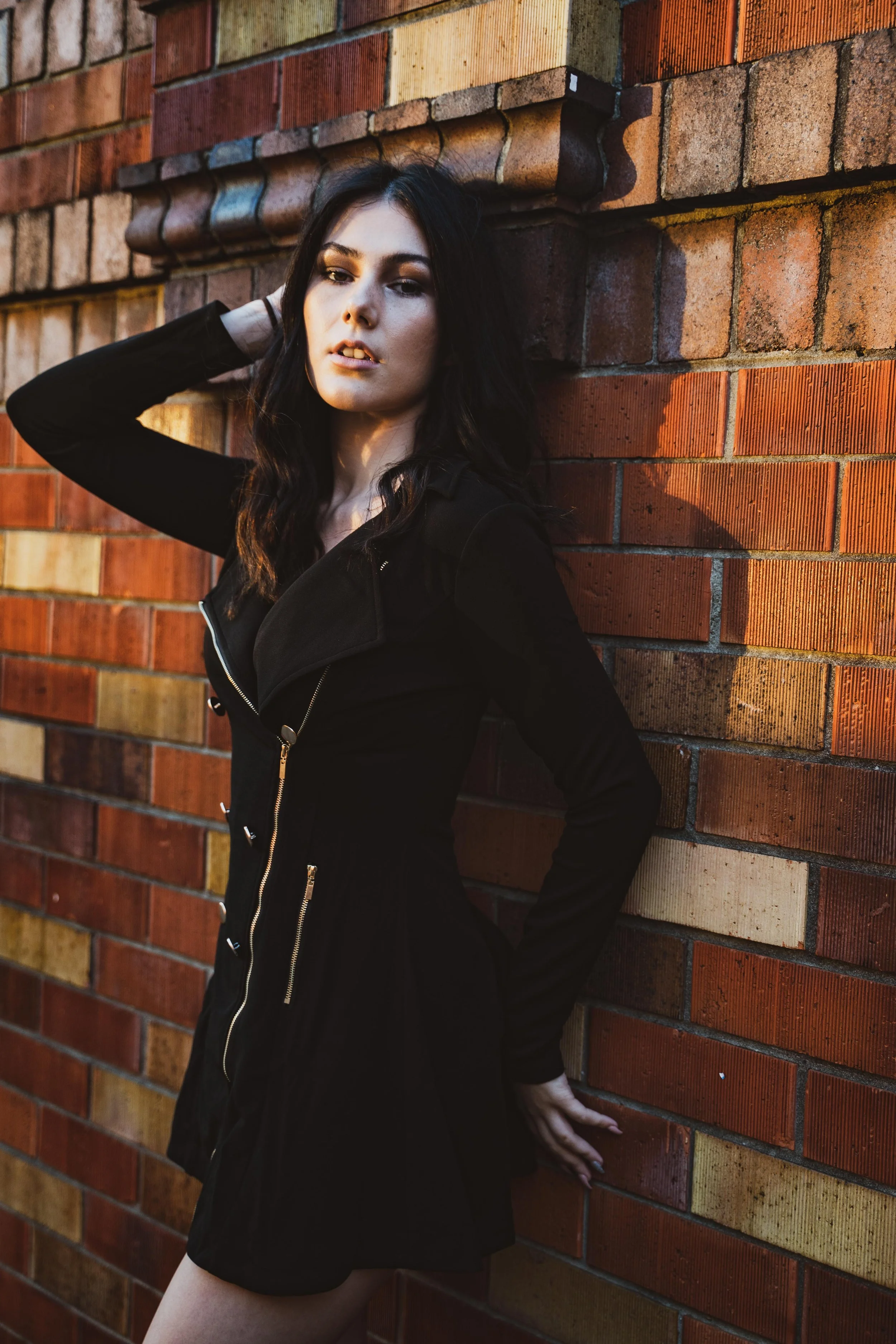 A woman with dark wavy hair and light skin leaning against a brick wall, wearing a black dress with zippers.