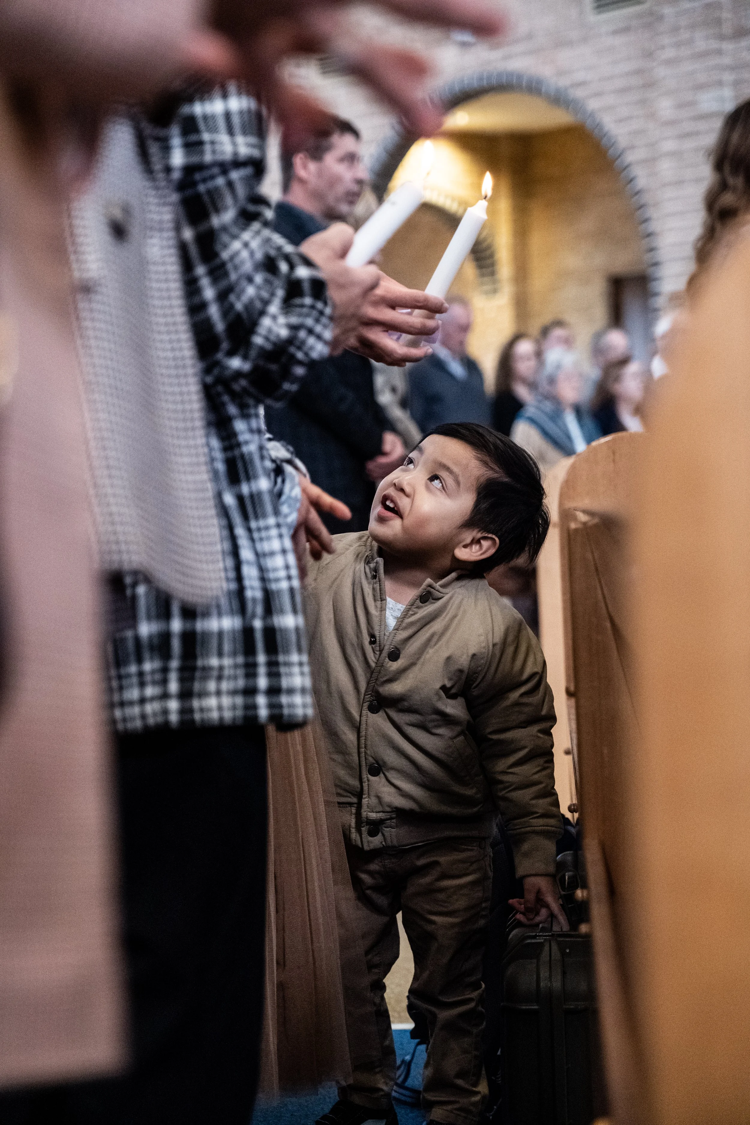 A young boy during a religious candle lighting ceremony in a church, looking up at an adult holding a lit candle, with other attendees in the background.