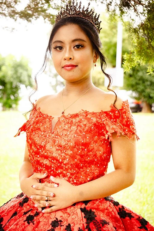 Young woman wearing a red lace dress with floral embroidery, a black tiara, and jewelry, standing outside with trees in the background.