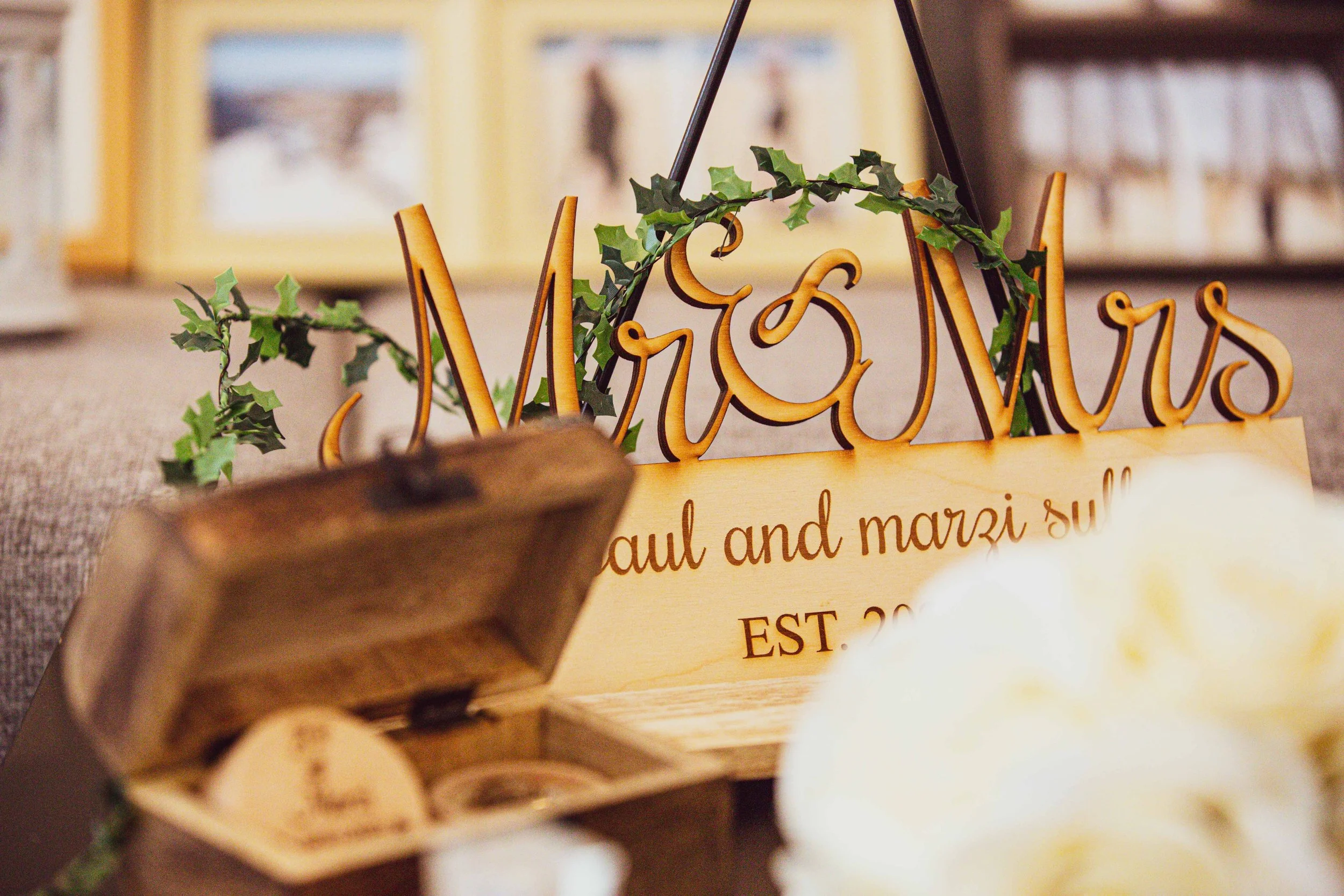 Wooden wedding sign that says 'Mrs. & Mrs.' with green ivy decorations, partially obscured text reading 'Paul and Marzi' and 'EST. 20', placed on a table with a small wooden box and blurred white flower in the foreground.
