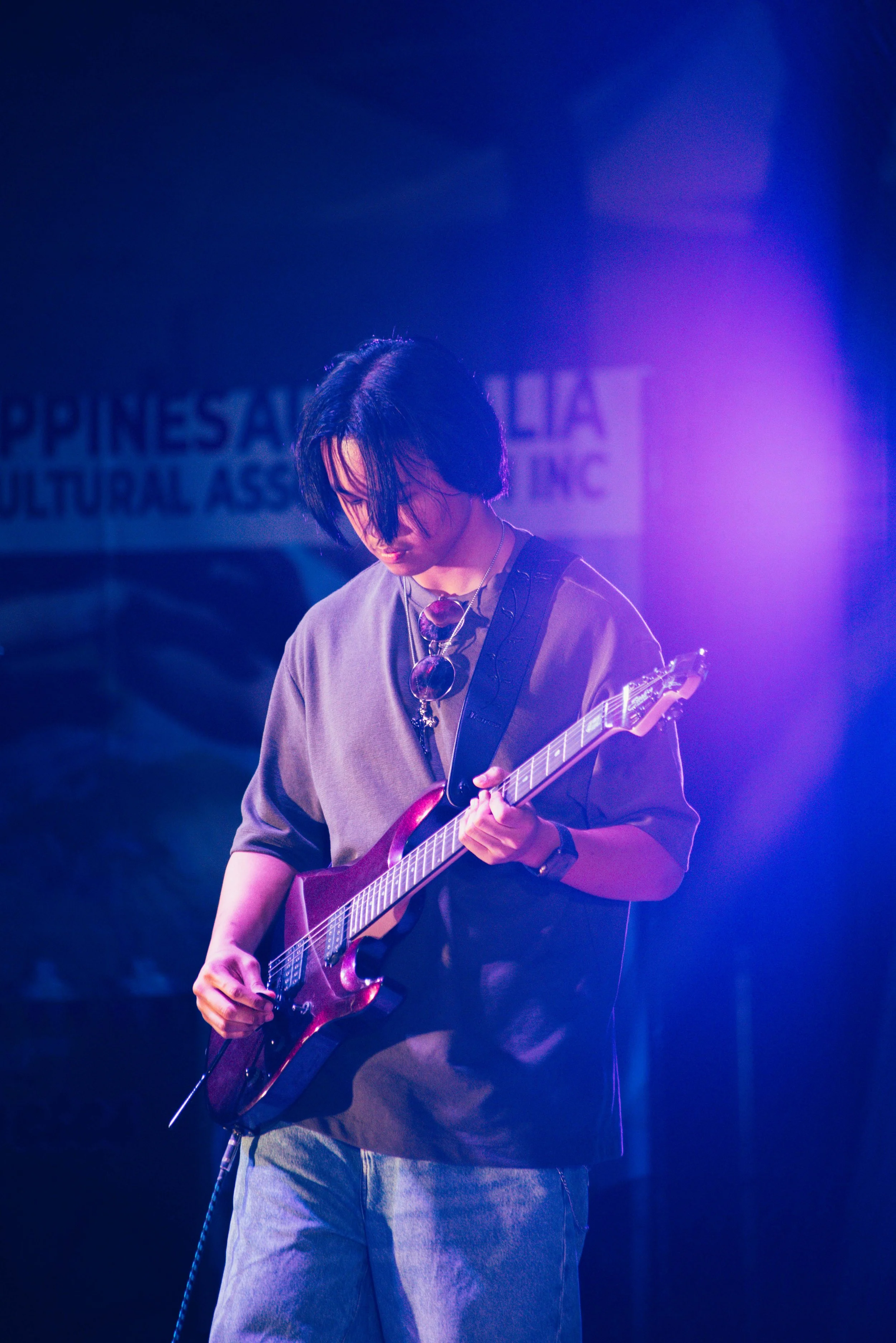 A young person with black hair playing a red electric guitar on stage under purple lighting, with a backdrop that reads 'Philippine Cultural Association'.