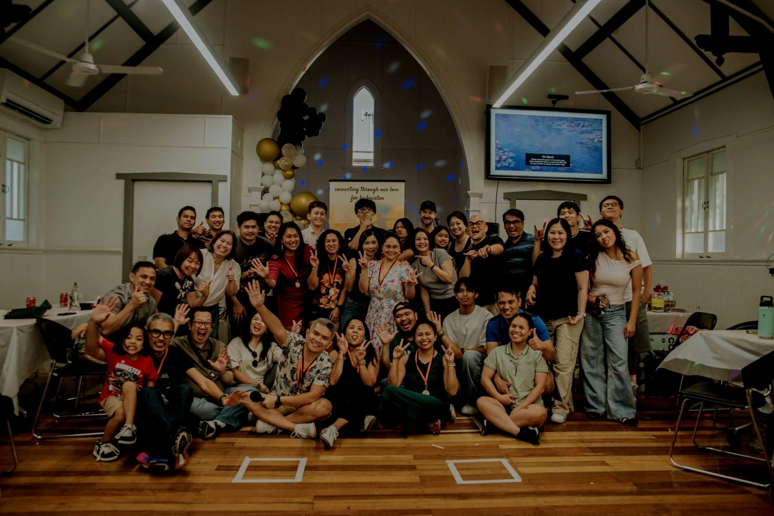 Group of people gathered indoors in a church or hall, smiling and posing for a group photo, with some raising their hands and making peace signs, in front of balloons and a banner.