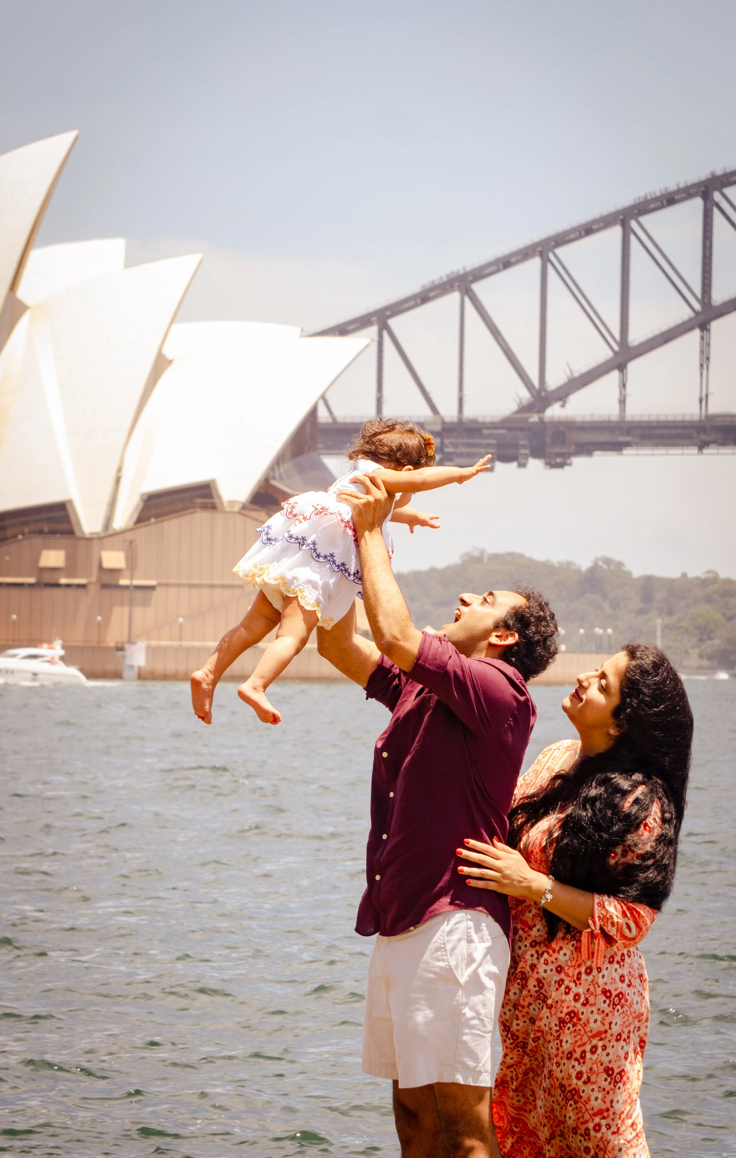 A family of three at the waterfront with the Sydney Opera House and Harbour Bridge in the background. The father lifts the young girl, and the mother looks on affectionately.