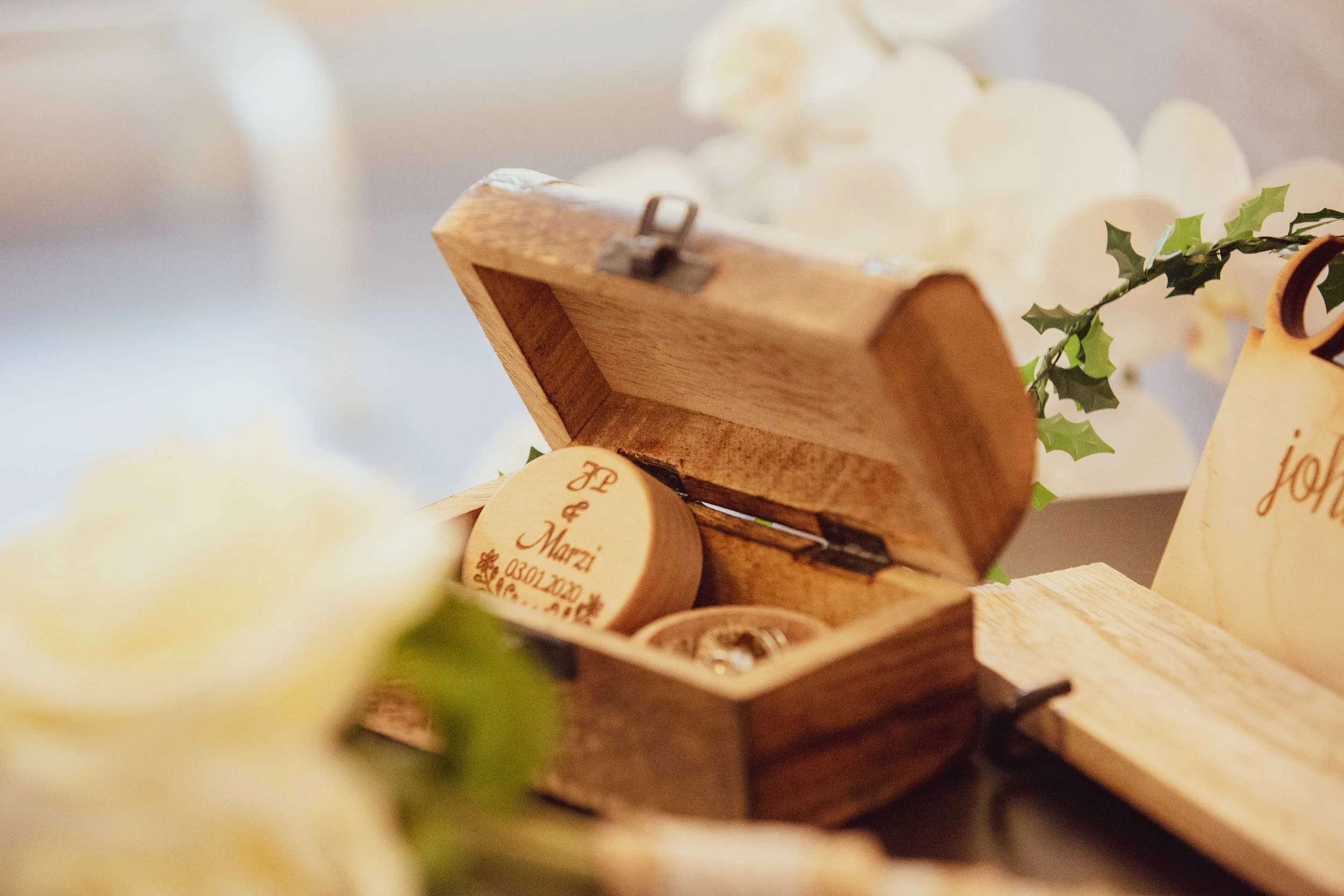 Wooden box with a lid open, containing a small round wooden keepsake with engraved text, surrounded by flowers and green holly leaves, likely part of a wedding or special event.