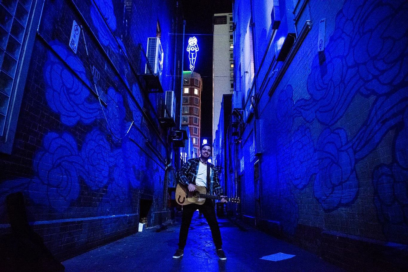 A man standing in a narrow alleyway at night, playing an acoustic guitar and smiling, with blue neon lights illuminating graffiti-covered brick walls on either side.