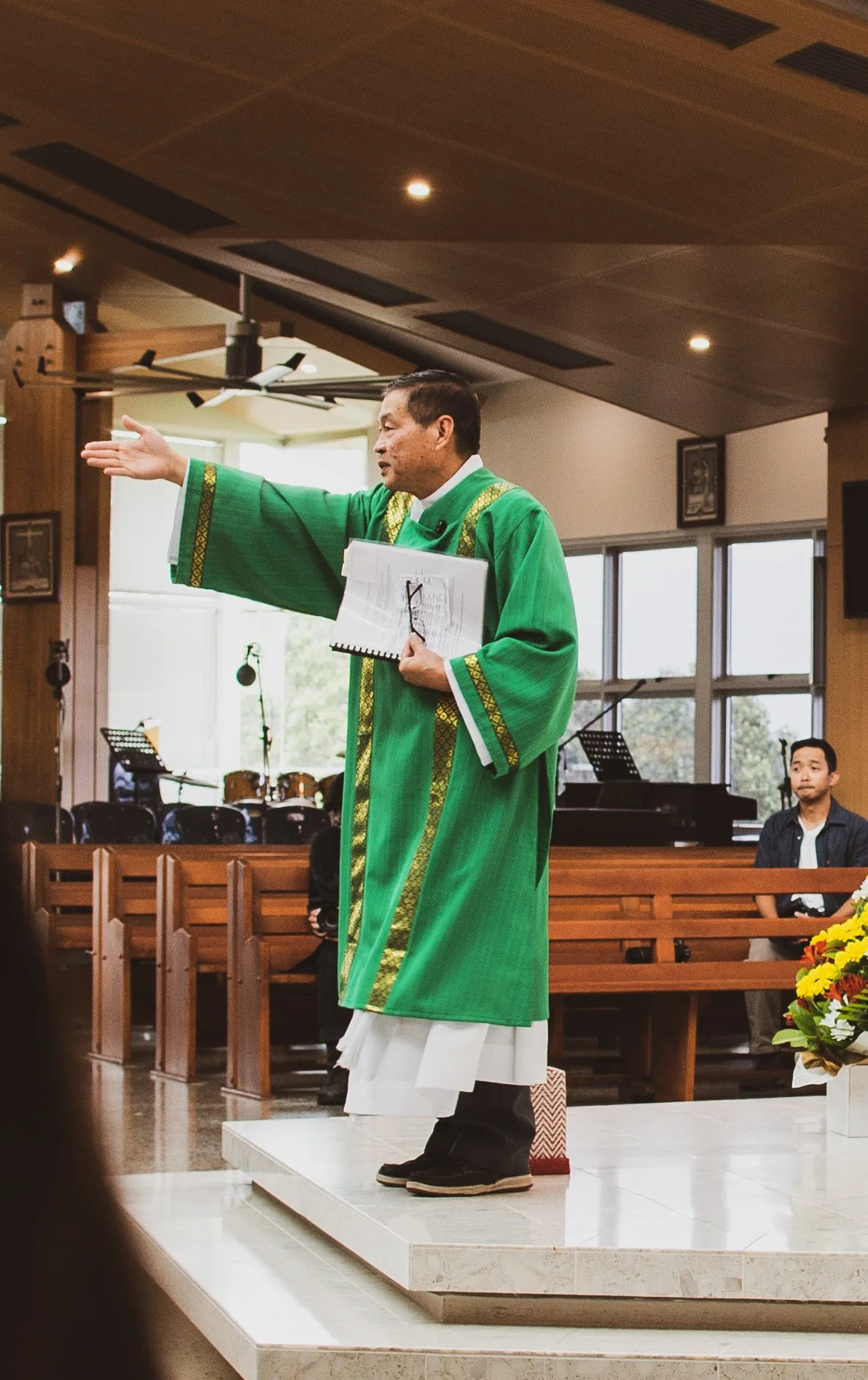 A priest in a green and gold liturgical robe stands on a stage in a church, gesturing with his right hand while holding a notebook and glasses in his left hand. There are musicians and a piano in the background, and a man seated in the pews to the ri