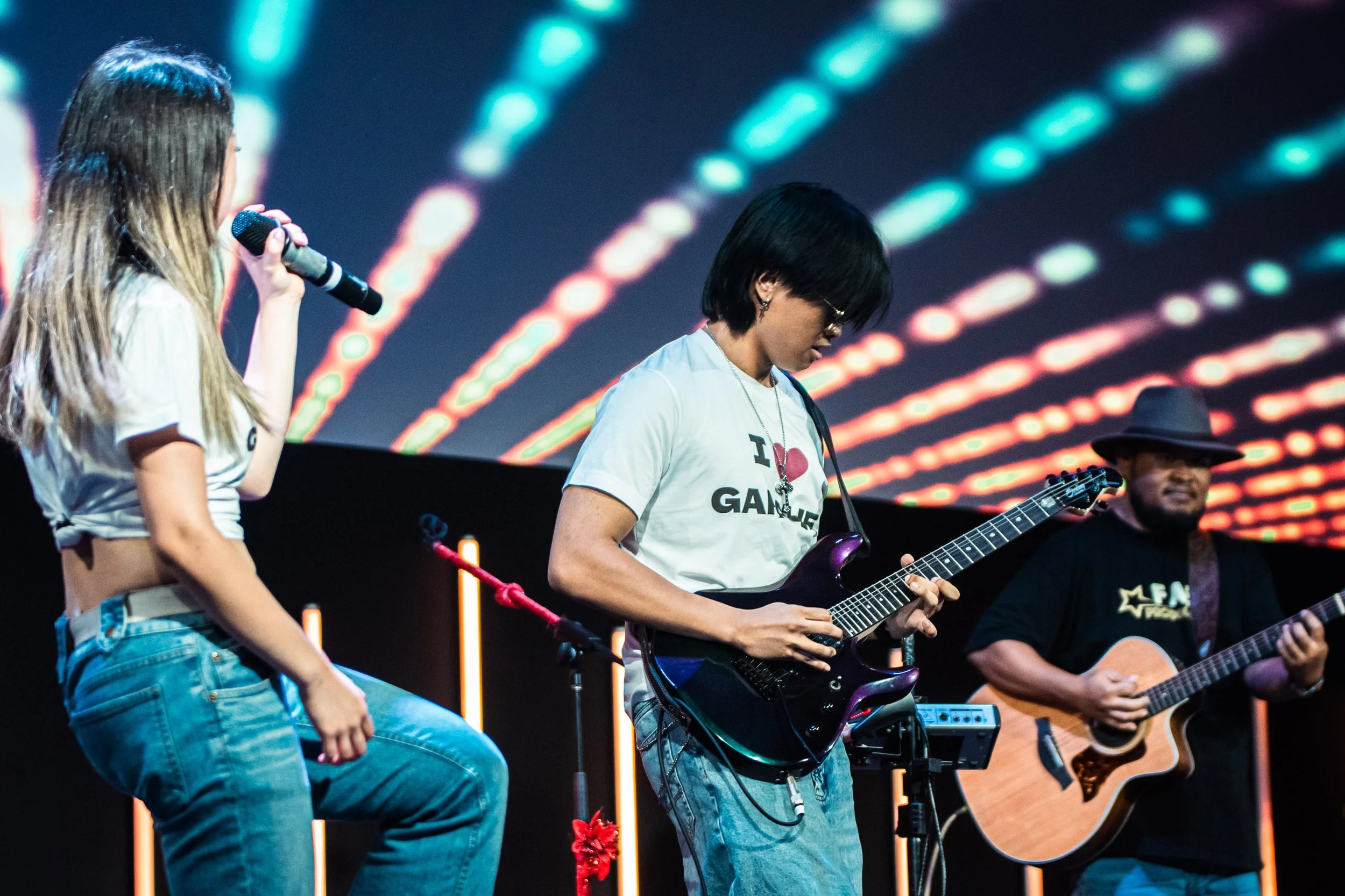 Three musicians performing on stage with colorful light patterns in the background: a young woman singing into a microphone, a woman playing an electric guitar, and a man playing an acoustic guitar.