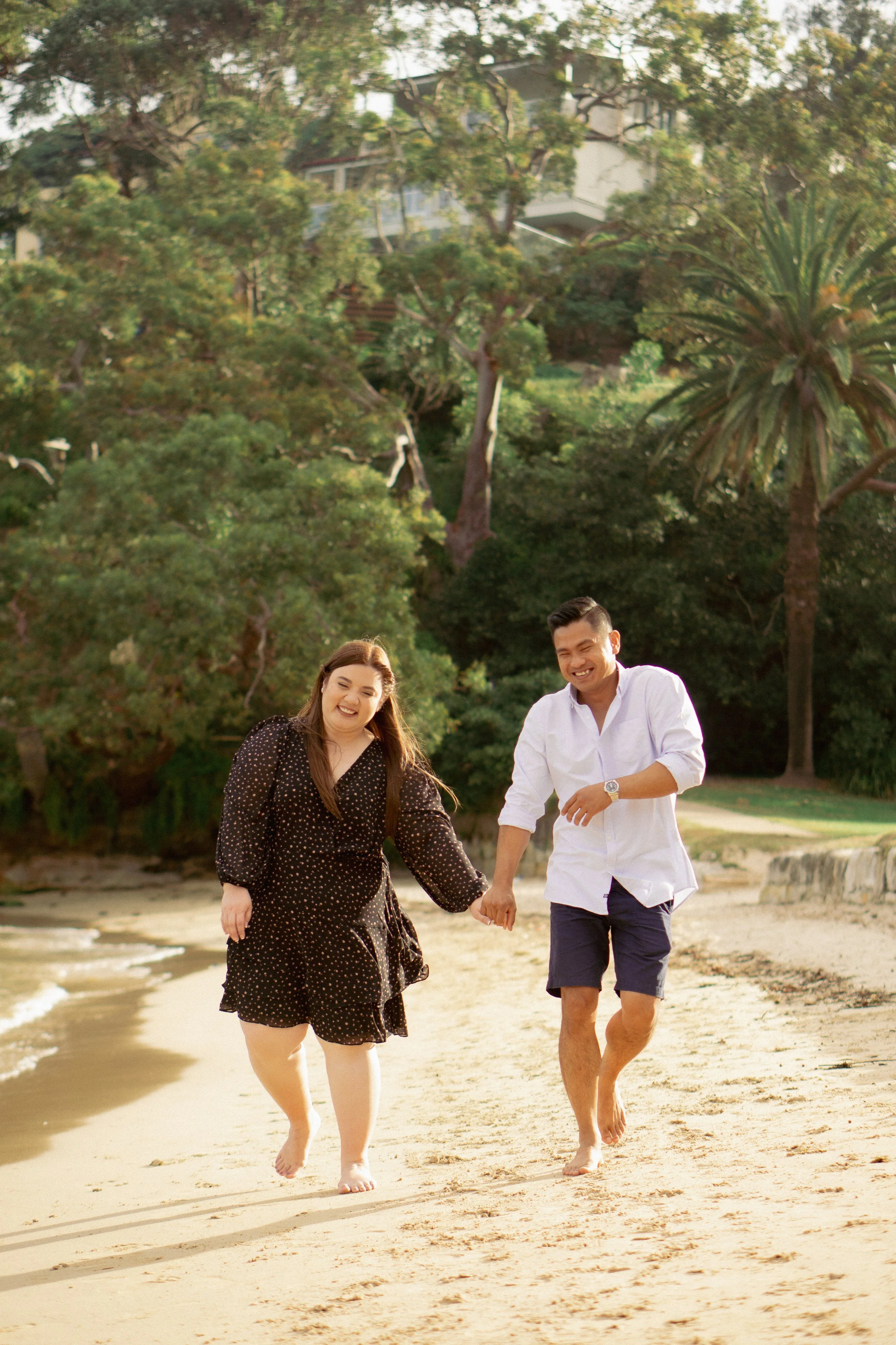 A couple holding hands and walking along the beach, smiling, with trees and houses in the background.