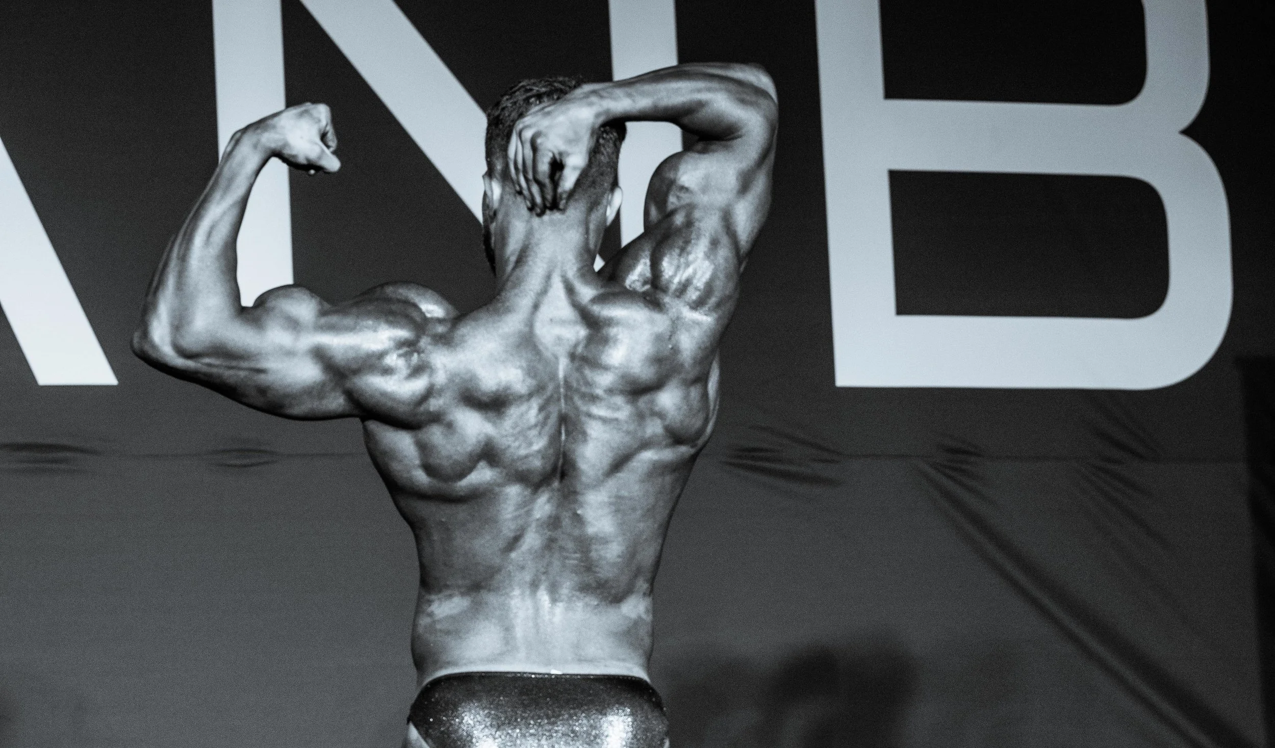 Black and white photo of a male bodybuilder with muscles flexed, facing away, on a stage with a background that has large letters.