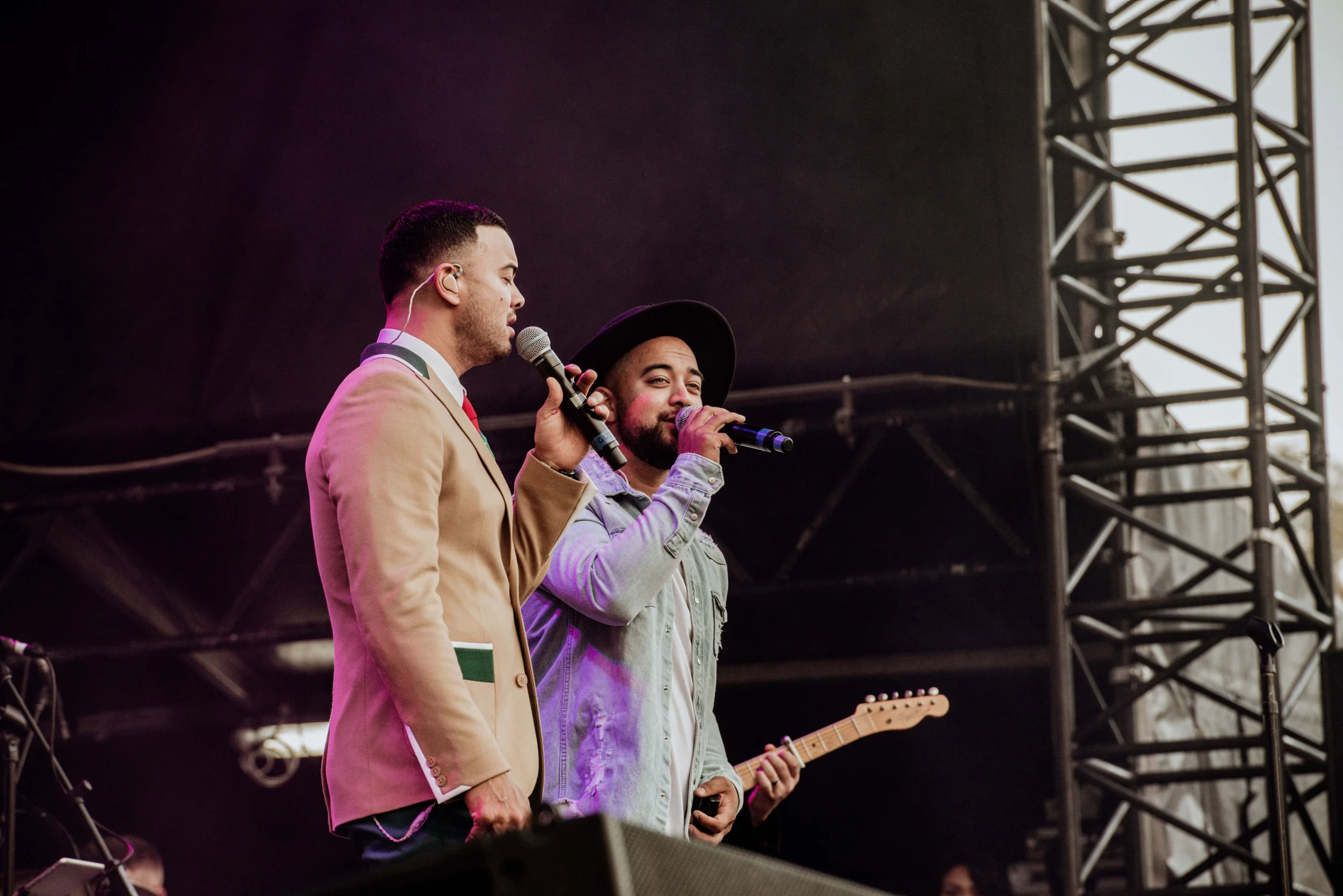 Two male singers holding microphones perform on stage, one wearing a beige blazer and the other in a denim jacket and black hat, with a guitarist partially visible in the background.