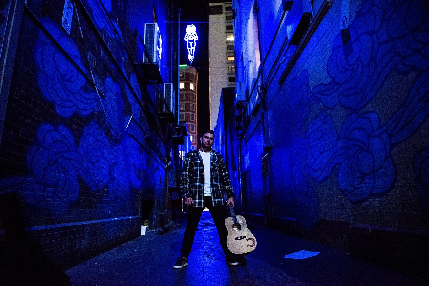 A man standing in a narrow alleyway illuminated by blue neon lights, holding an acoustic guitar, with blue mural artwork on the brick walls and city buildings visible in the background.