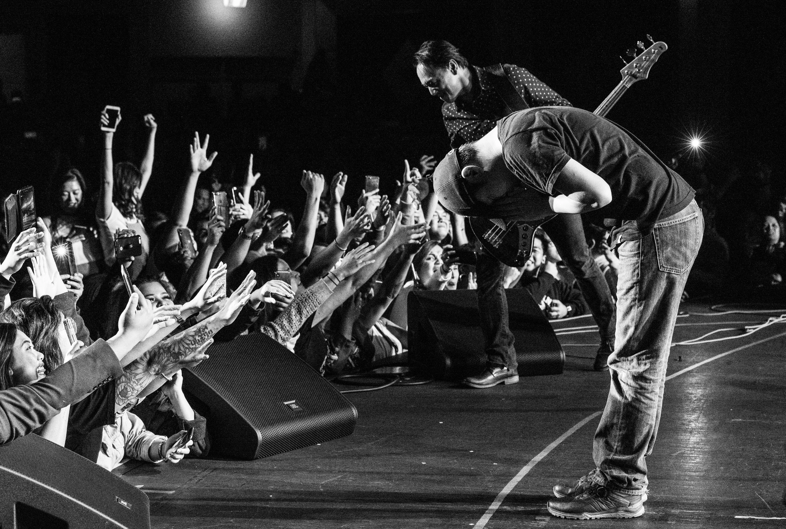 A black and white photo of a live music performance showing a man with a guitar bowing to the crowd on stage, with fans reaching out their hands, some holding phones, in front of him.
