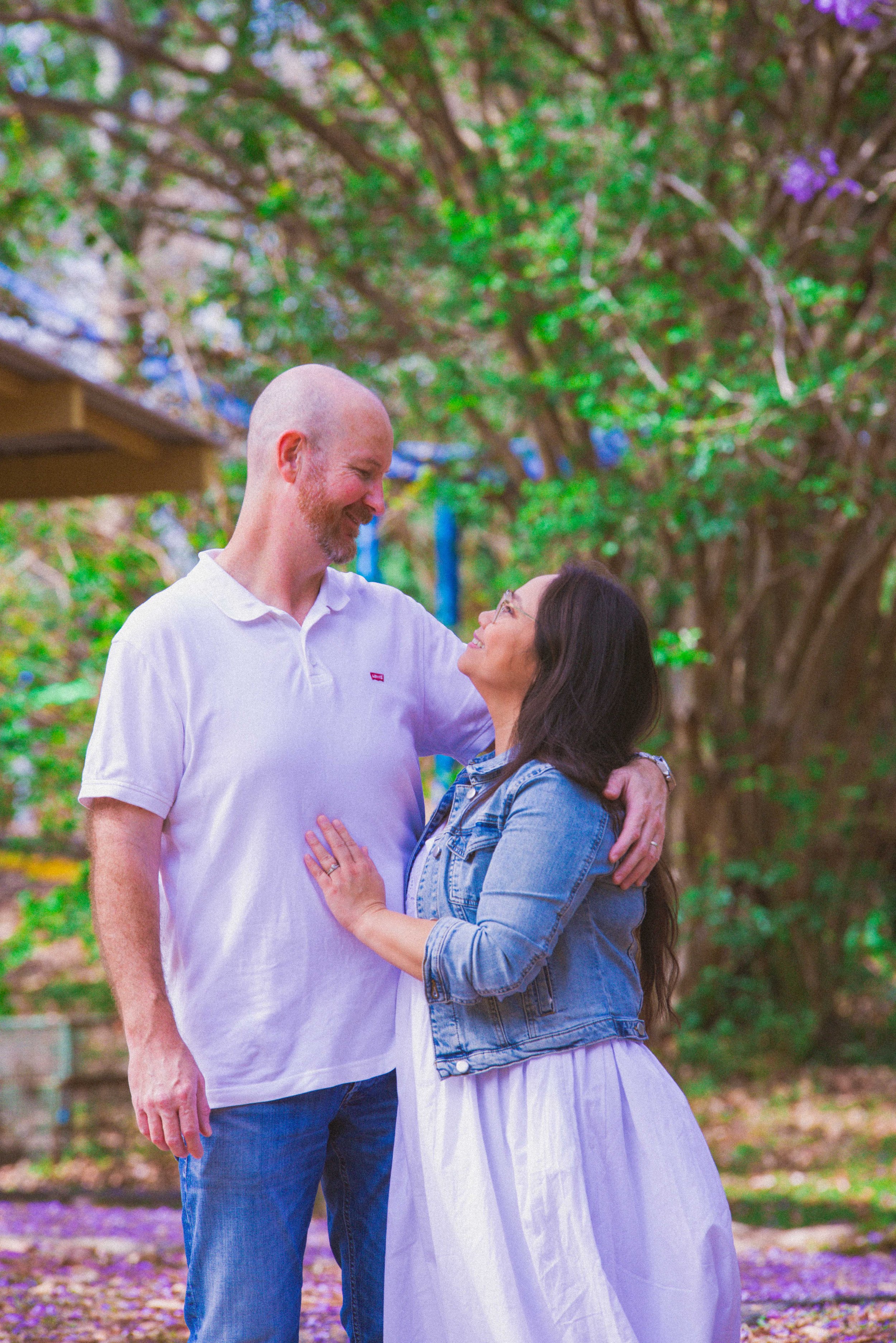 A man and woman standing close together outdoors, smiling and looking into each other's eyes, with trees and purple flowers in the background.