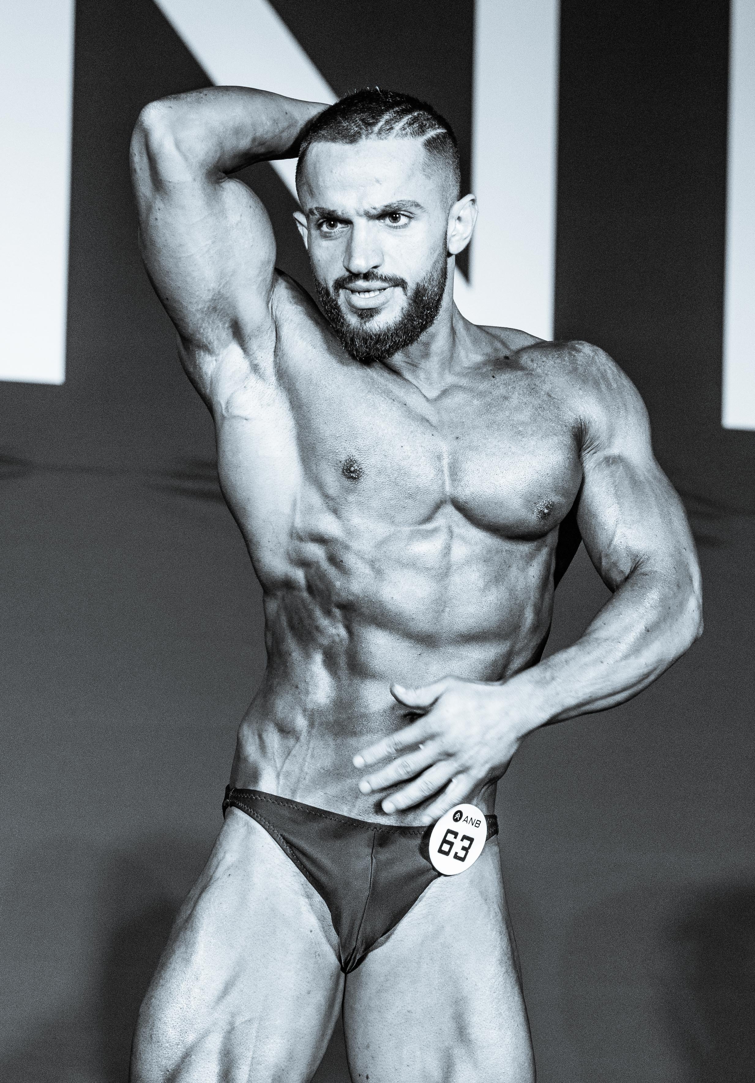 A male bodybuilder posing on stage during a competition, wearing a competitor number 63, with a muscular physique and a beard.