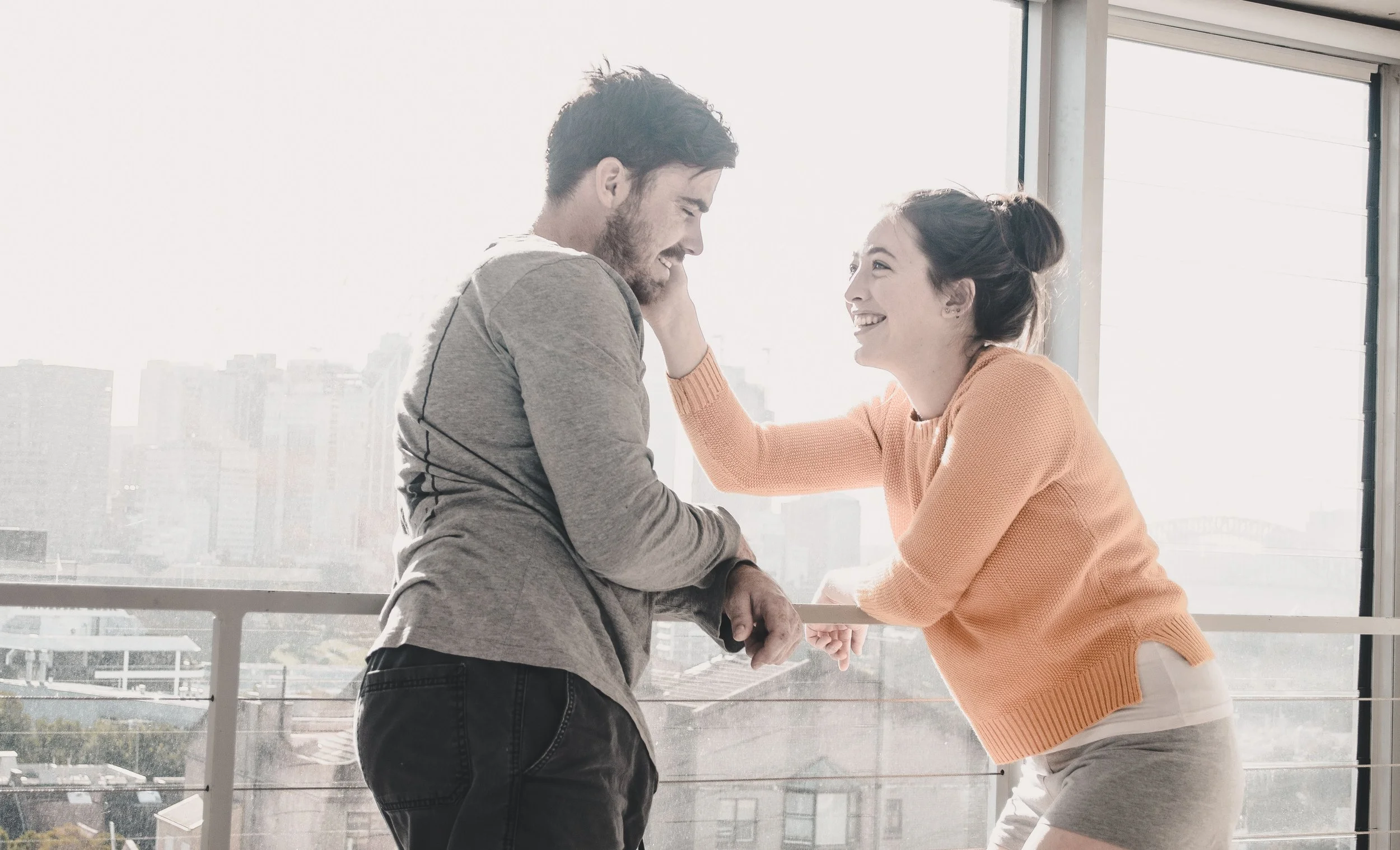 A smiling woman in an orange sweater and gray shorts playfully touching the face of a man in a gray hoodie, with a city skyline in the background.