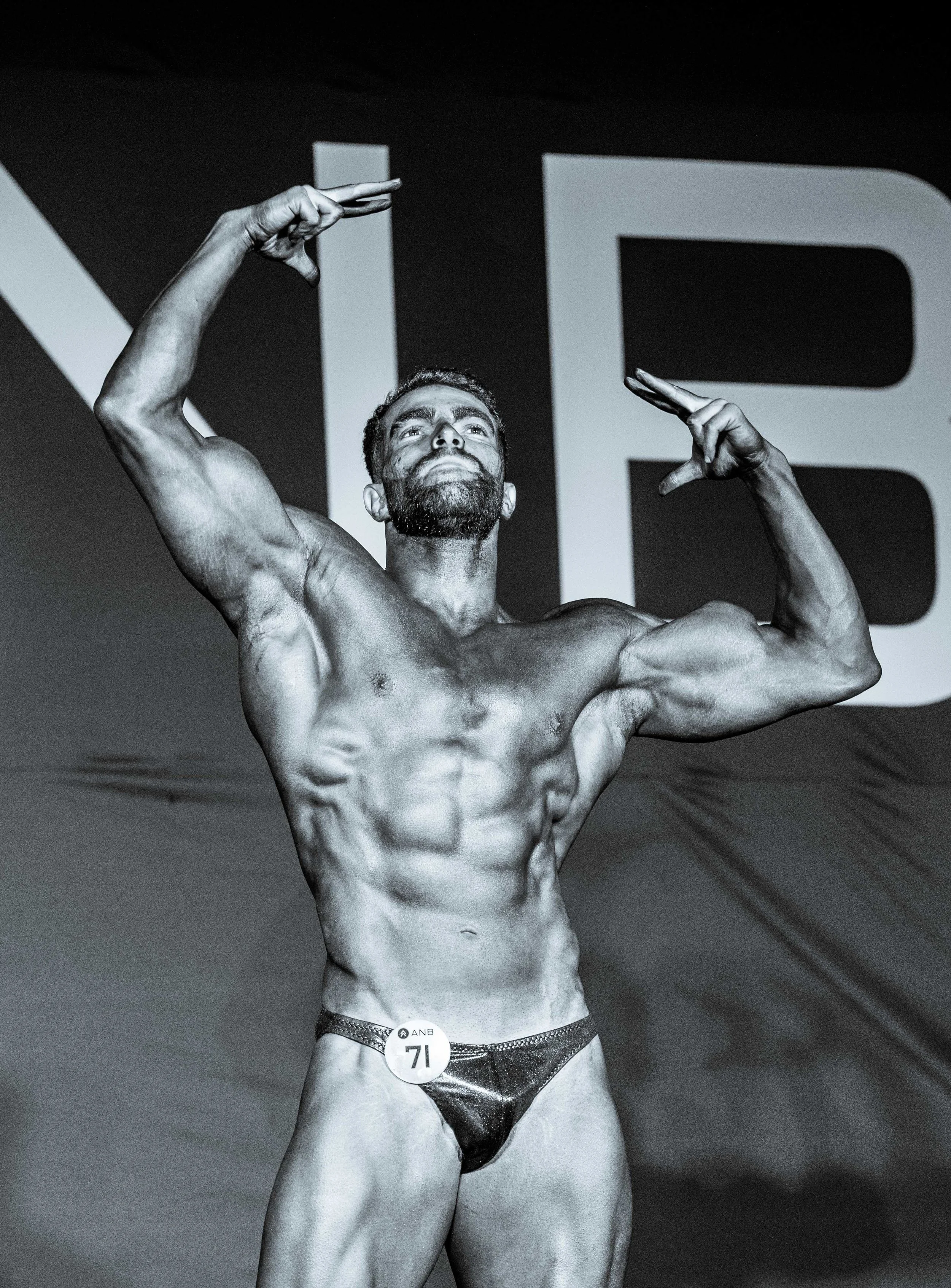 Black and white image of a male bodybuilder on stage, posing with flexed muscles, wearing a small black posing suit and a badge numbered '71'. Large letters are visible in the background.