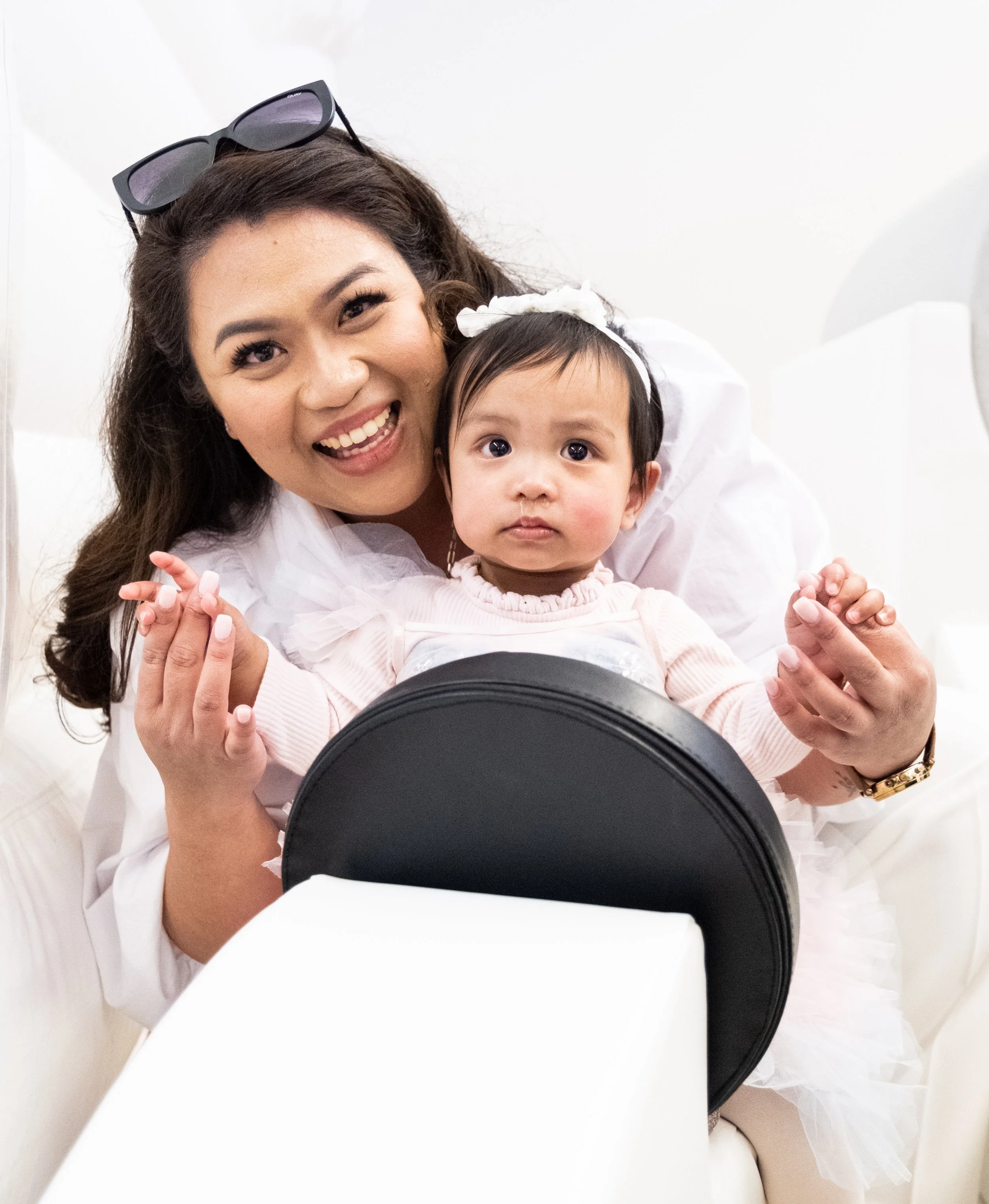 A smiling woman with long dark hair, wearing sunglasses on her head, is holding a young girl with short dark hair, dressed in light pink, in a white dress with frills. They are sitting together, facing the camera, with the woman's arms around the gir