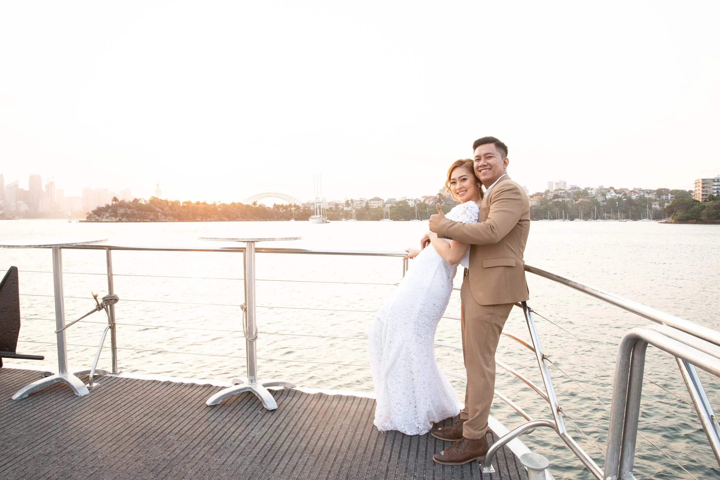 A happy couple in wedding attire on a boat, embracing and smiling at the camera, with a city skyline, water, and sailboats in the background during sunset.