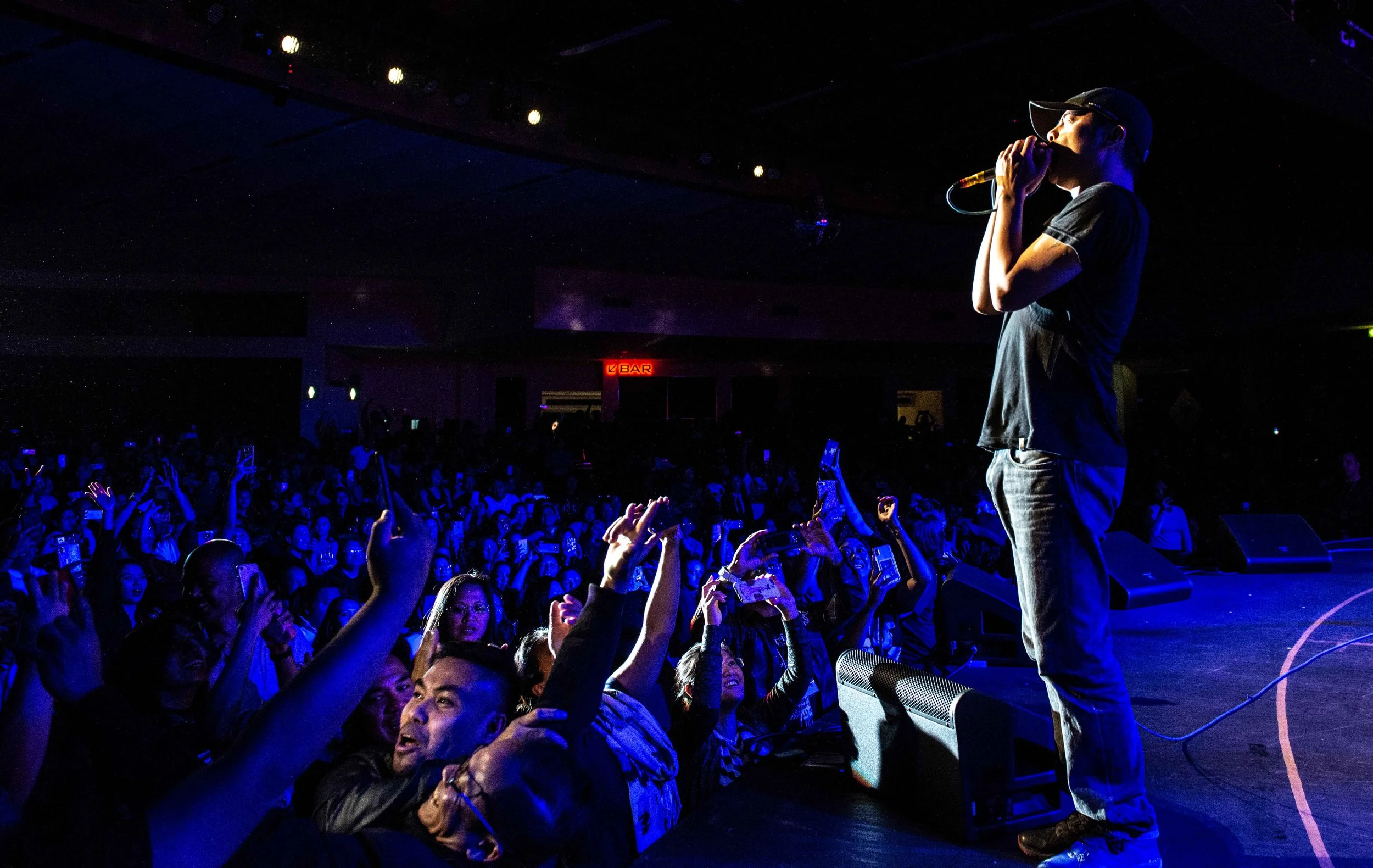 Performer on stage singing into microphone at concert with audience raising hands and taking photos in the dark