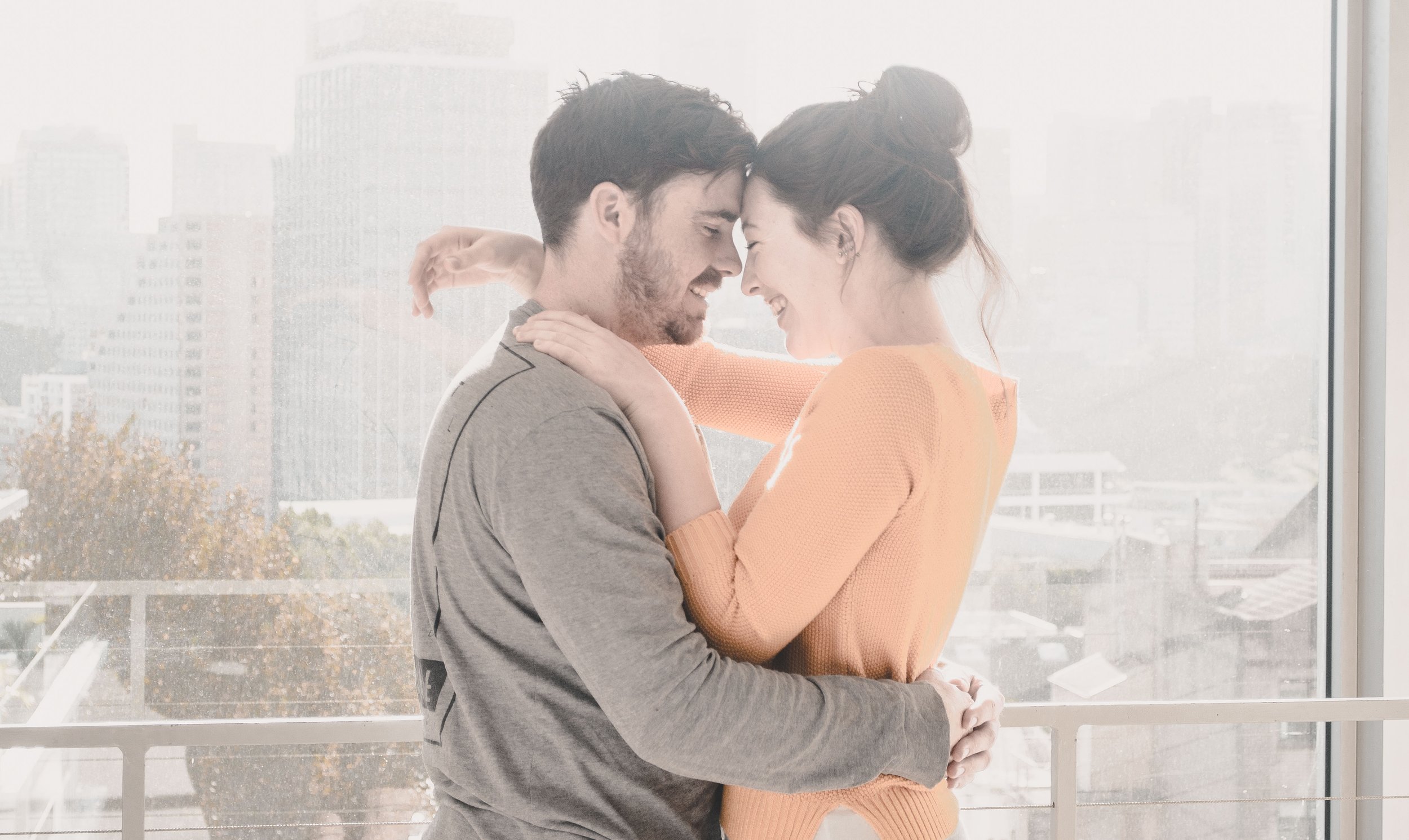 A young couple standing close together indoors by a large window, smiling and touching foreheads, with a cityscape in the background.