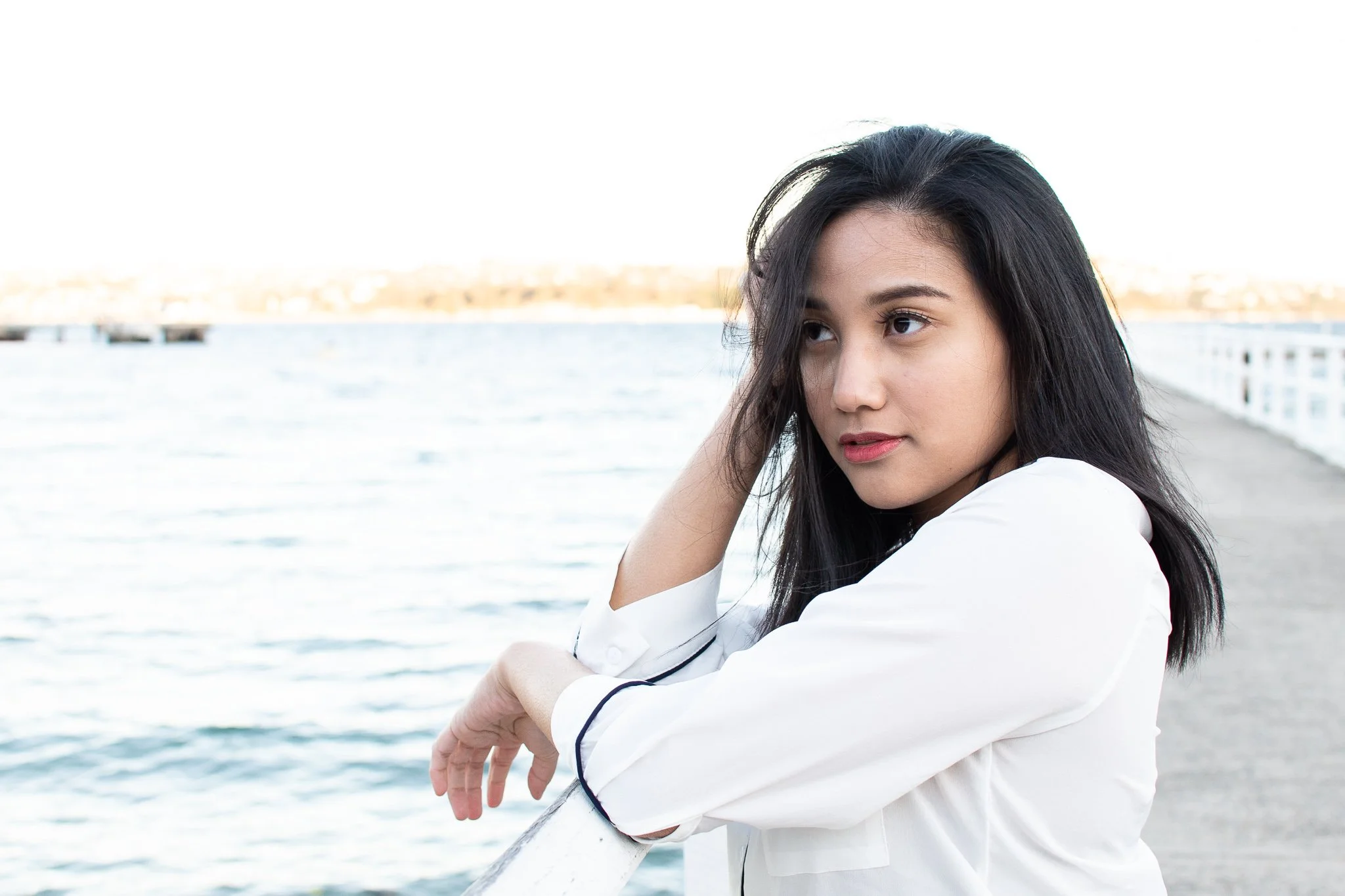 Young woman with black hair leaning on a railing by a body of water, wearing a white shirt, with a distant shoreline in the background.