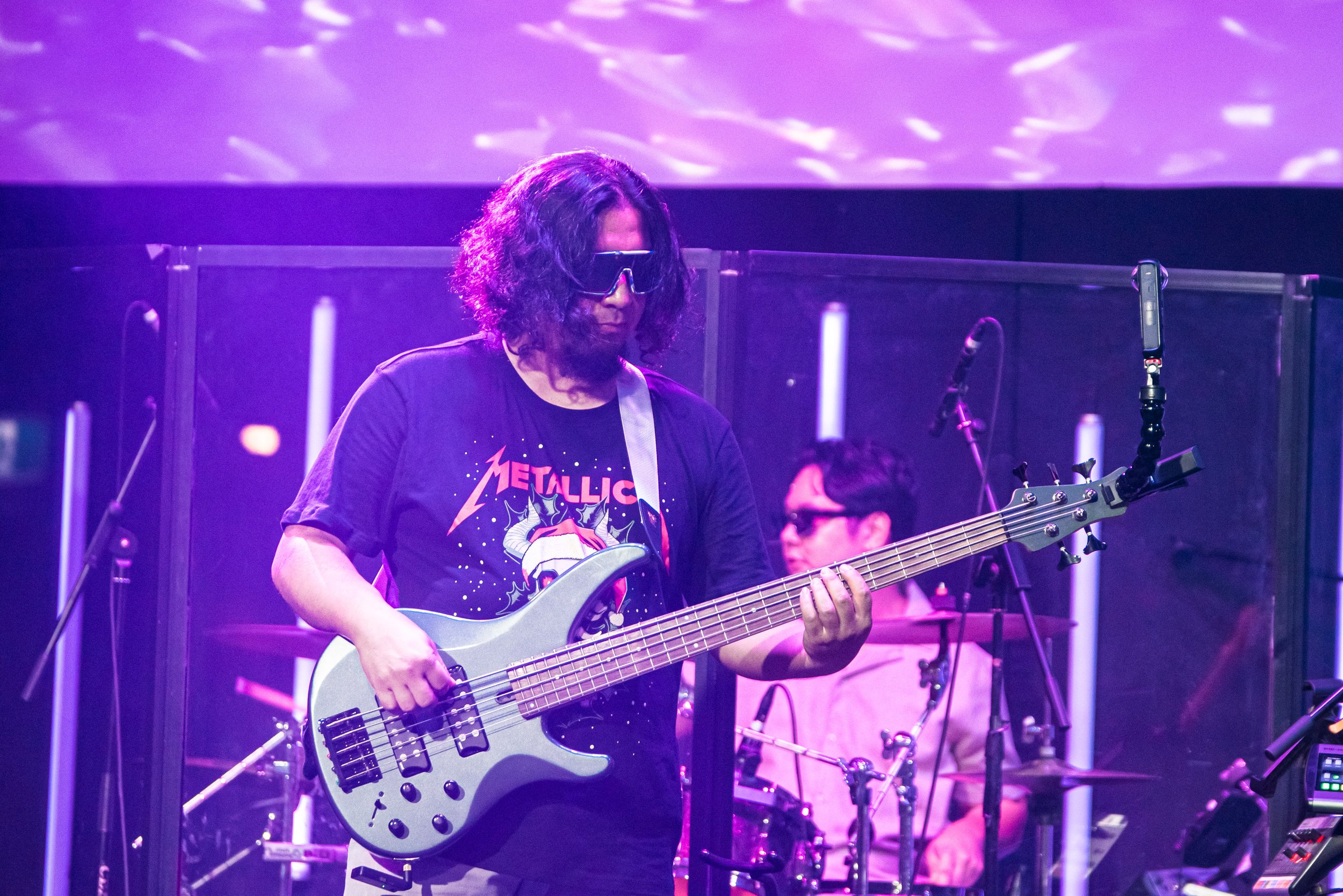 Musician playing a bass guitar on stage with purple stage lighting, wearing a Black Metallica T-shirt and sunglasses, with a drummer in the background.