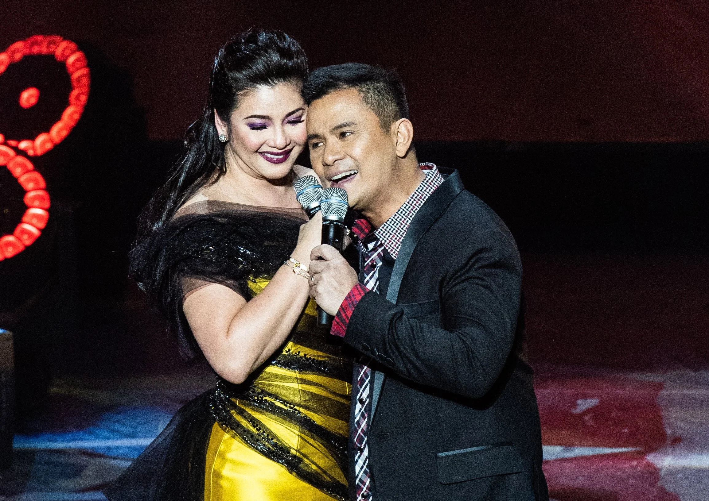 A woman in a yellow and black dress and a man in a black blazer are singing together with microphones, smiling and close to each other, on stage with a dark background.