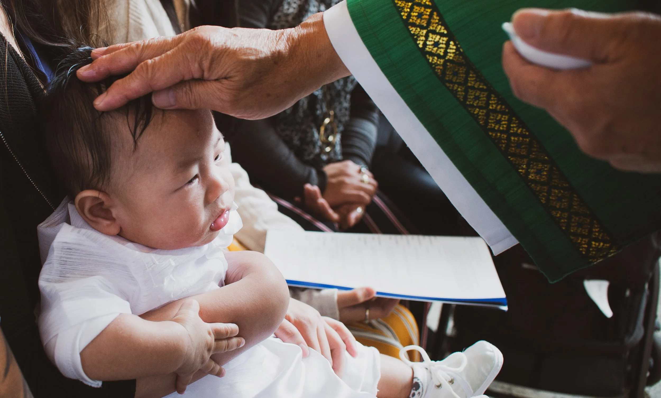 A young child sitting in a wheelchair, receiving a blessing or prayer from an elderly person who is touching their head. The elderly person holds a document or book.