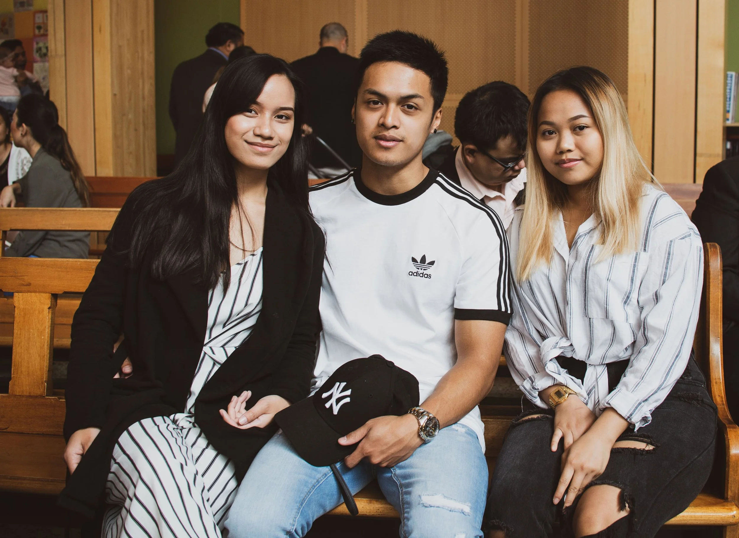Three young adults, two women and one man, sitting together on a wooden bench indoors, looking at the camera, with other people in the background.